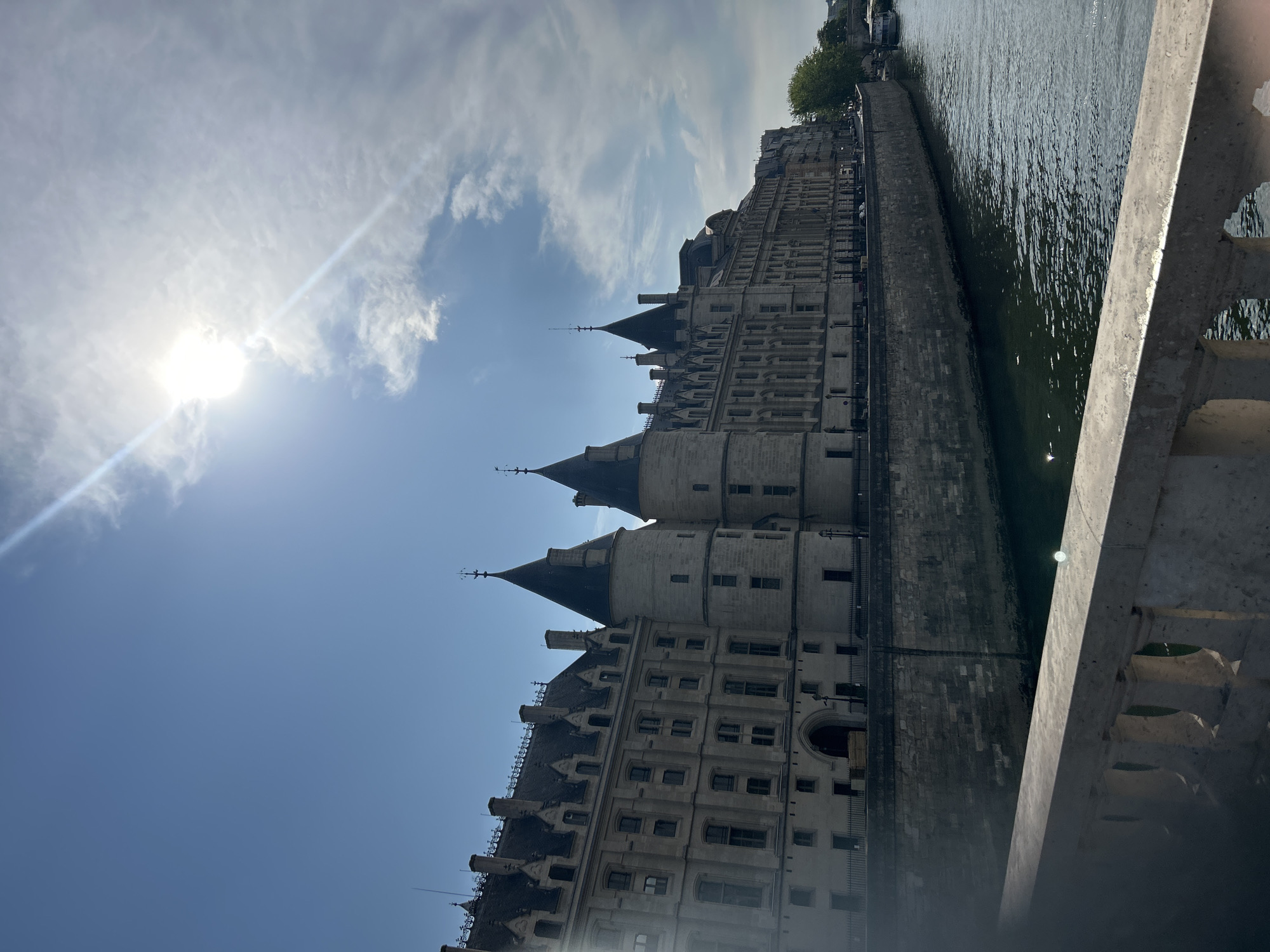 The image shows the Conciergerie on the Île de la Cité in Paris, France, viewed from a bridge. The building features Gothic architecture with conical turrets and tall windows. A stone embankment separates the building from the dark, rippling water of the Seine River. A light-colored stone railing from the bridge is visible in the foreground. The sky is blue with some white clouds and the sun is shining brightly, creating a lens flare effect. A boat is visible on the river in the distance.