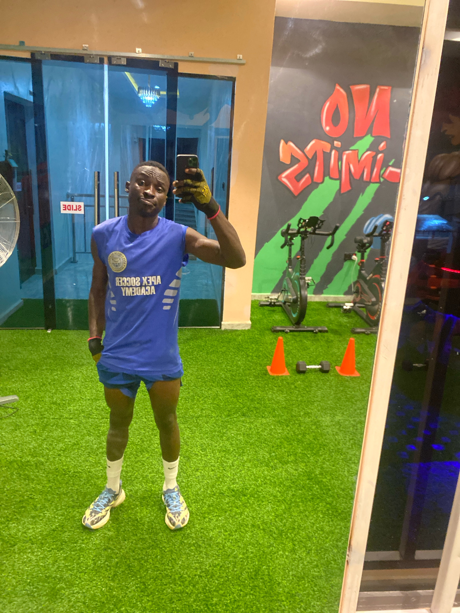 The image shows a young Black male posing for a selfie in a gym. He is wearing a blue sleeveless t-shirt with "APEX SOCCER ACADEMY" printed on it in white lettering, blue athletic shorts, white socks, and patterned athletic shoes. He is holding a black smartphone in his right hand, wearing a yellow and black glove on his right hand, and a red wristband on his left wrist. The gym has bright green artificial turf flooring. In the background, there are large glass doors with "SLIDE" written in red text, two stationary bicycles, orange traffic cones, and a dumbbell. A wall behind the bicycles has graffiti that reads "NO LIMITS" in red and green lettering. The overall lighting suggests it is indoors, possibly at night. The specific location in Barden Yana, Nigeria is not discernible from the image itself.