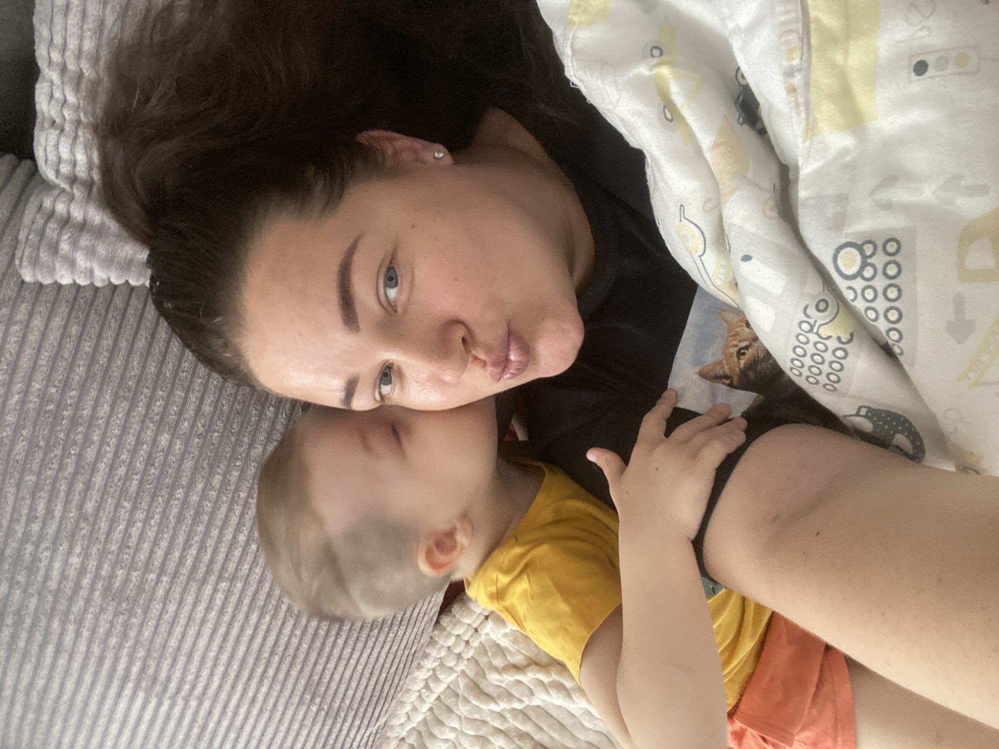 A woman and a young child are lying on a bed. The woman has dark hair, fair skin, and blue eyes. She is wearing a black shirt. The child has light hair and is wearing a yellow shirt and orange shorts. The child is kissing the woman on the cheek. There is a blanket with a yellow and grey pattern over them. A cat is peering out from under the blanket. The bed has a grey headboard. The location is not identifiable as Sochi, Russia.