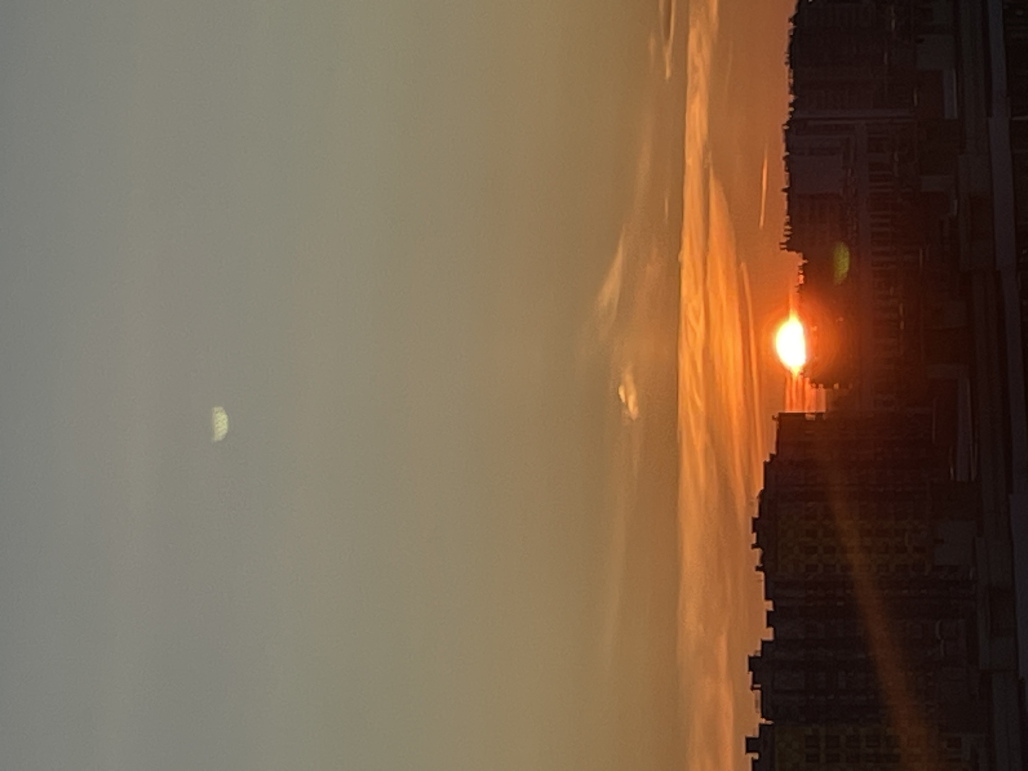 The image shows a sunset over a city. The sky is a gradient of orange and yellow, with some wispy clouds. The sun is a bright orange disc, low on the horizon, partially obscured by buildings. Silhouettes of apartment buildings fill the lower portion of the image, with some windows appearing to glow from interior lights. There is a faint, crescent-shaped object visible in the upper portion of the sky, which could be the moon or a lens flare. The overall impression is a peaceful, urban sunset.