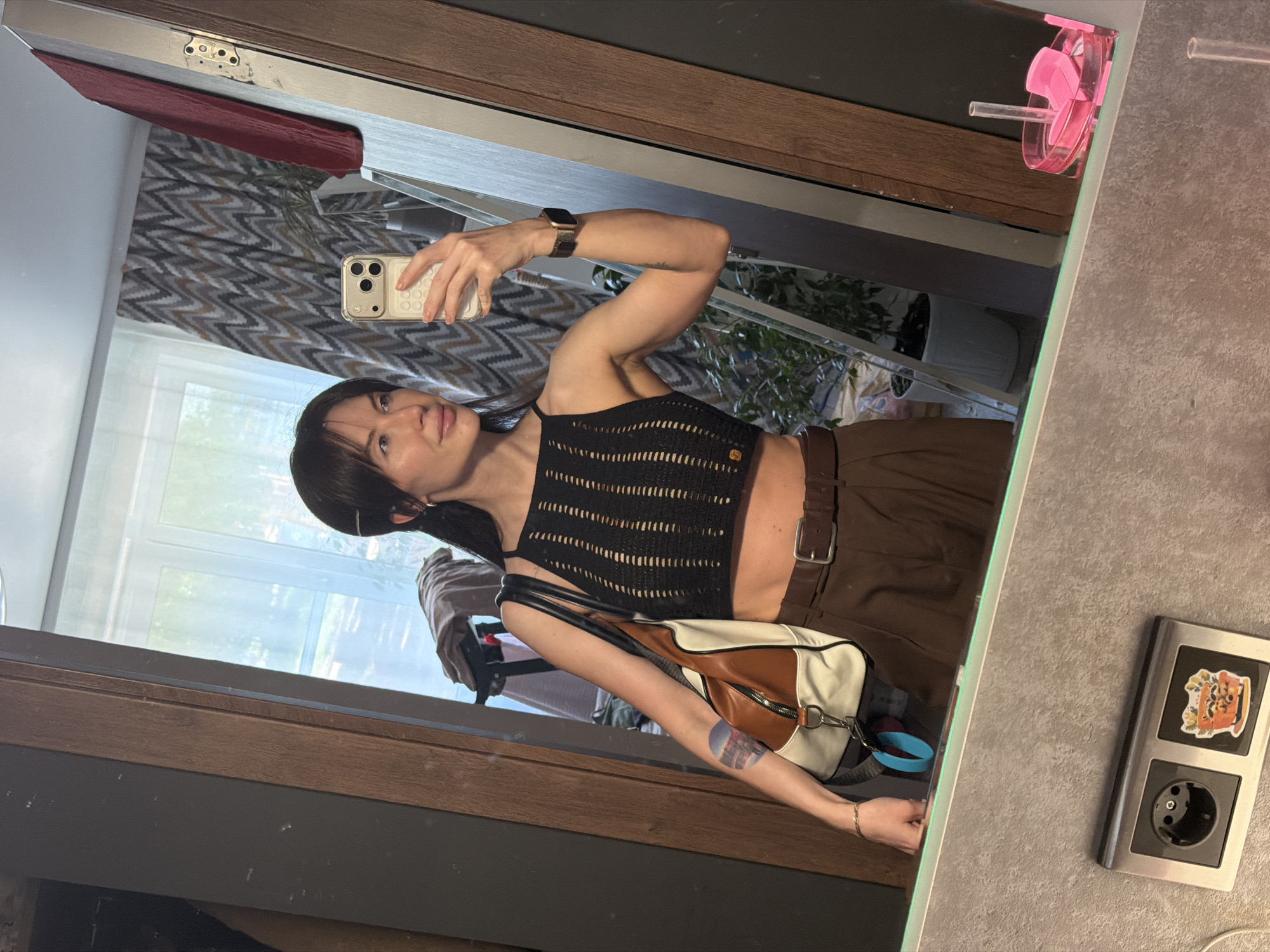 A woman stands in front of a mirror, holding a smartphone in her right hand to take a selfie. She is wearing a black, sleeveless top and brown trousers with a belt. A shoulder bag hangs from her left shoulder. The mirror reflects a room with a window, patterned curtains, and plants. To the right of the mirror is a light gray wall with a power outlet featuring a sticker. The image appears to be taken indoors in a residential setting. The city mentioned, Perm, Russia, is not directly discernable from the image's contents.