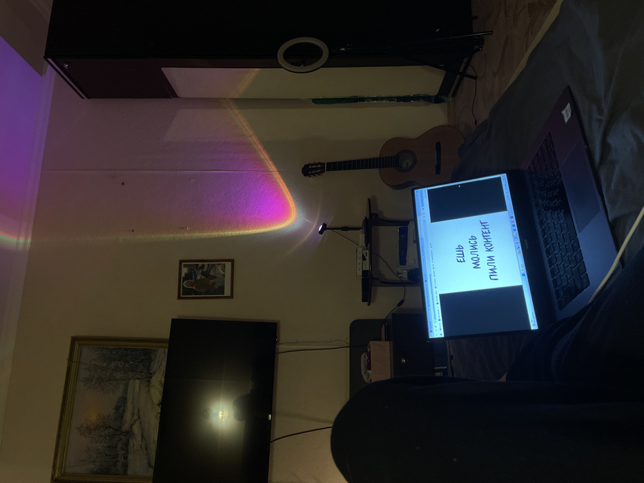 A room contains a laptop displaying text in Russian, "ЕШЬ МОЛИСЬ ПИЛИ КОНТЕНТ". A guitar hangs on the wall above a TV. A rainbow projection emanates from a device illuminating a portion of the wall. Other wall decor includes framed art. A ring light is visible attached to a high cabinet. The person is sitting on the bed with the laptop and phone in their lap.