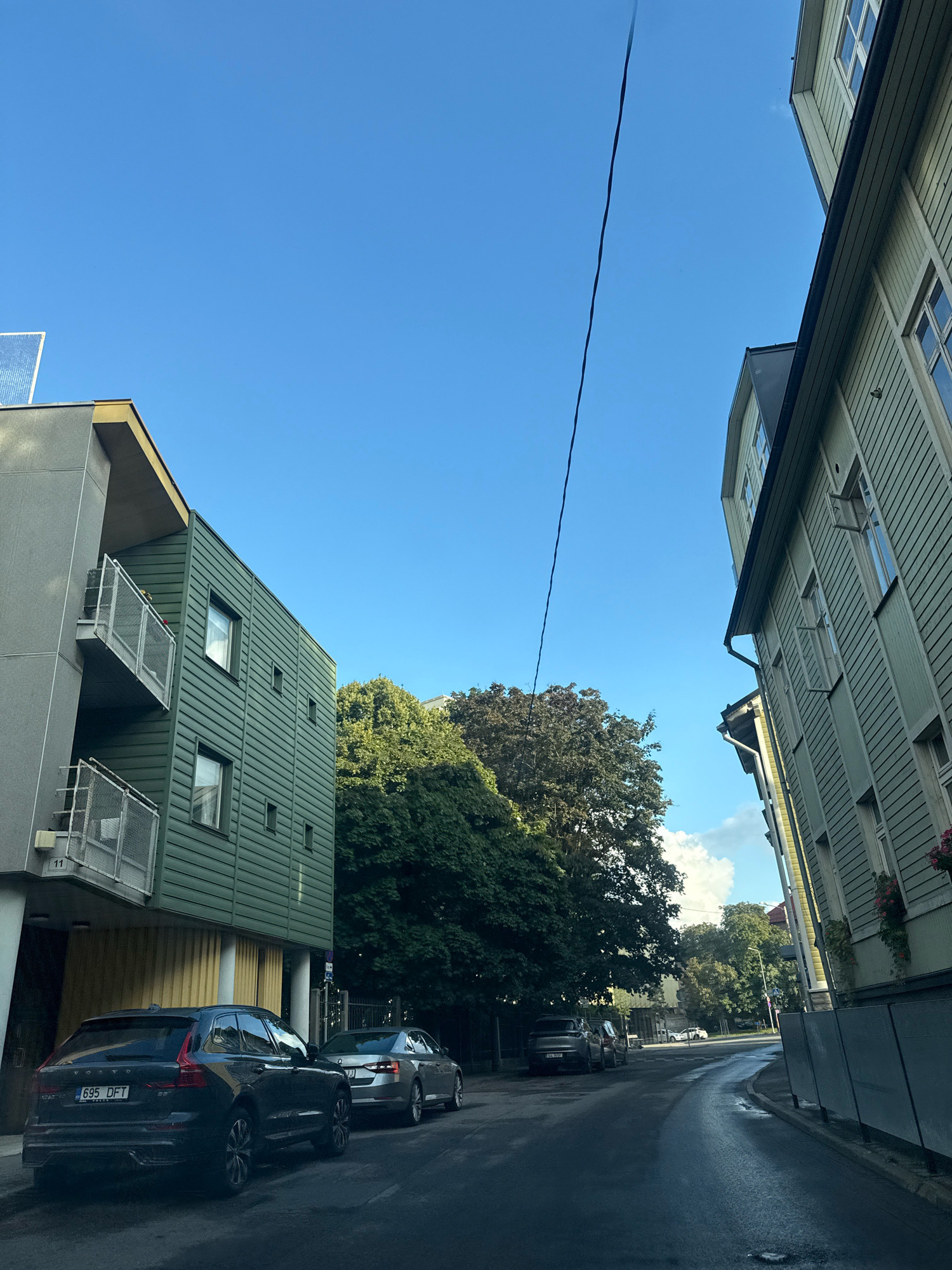 The image shows a street scene in Tallinn, Estonia. On the left is a modern building with grey and green siding, featuring balconies and windows. To the right is a traditional wooden building painted light green, with several windows, some open, and white trim. A power line stretches diagonally across the bright blue sky. Several cars are parked on the side of the wet asphalt road. The foreground shows the rear of a dark SUV with license plate "695 DFT" parked on the left, and a silver sedan to its right. Further down the street, more cars are visible, and trees line the sidewalk. The overall impression is an urban residential street on a clear day.