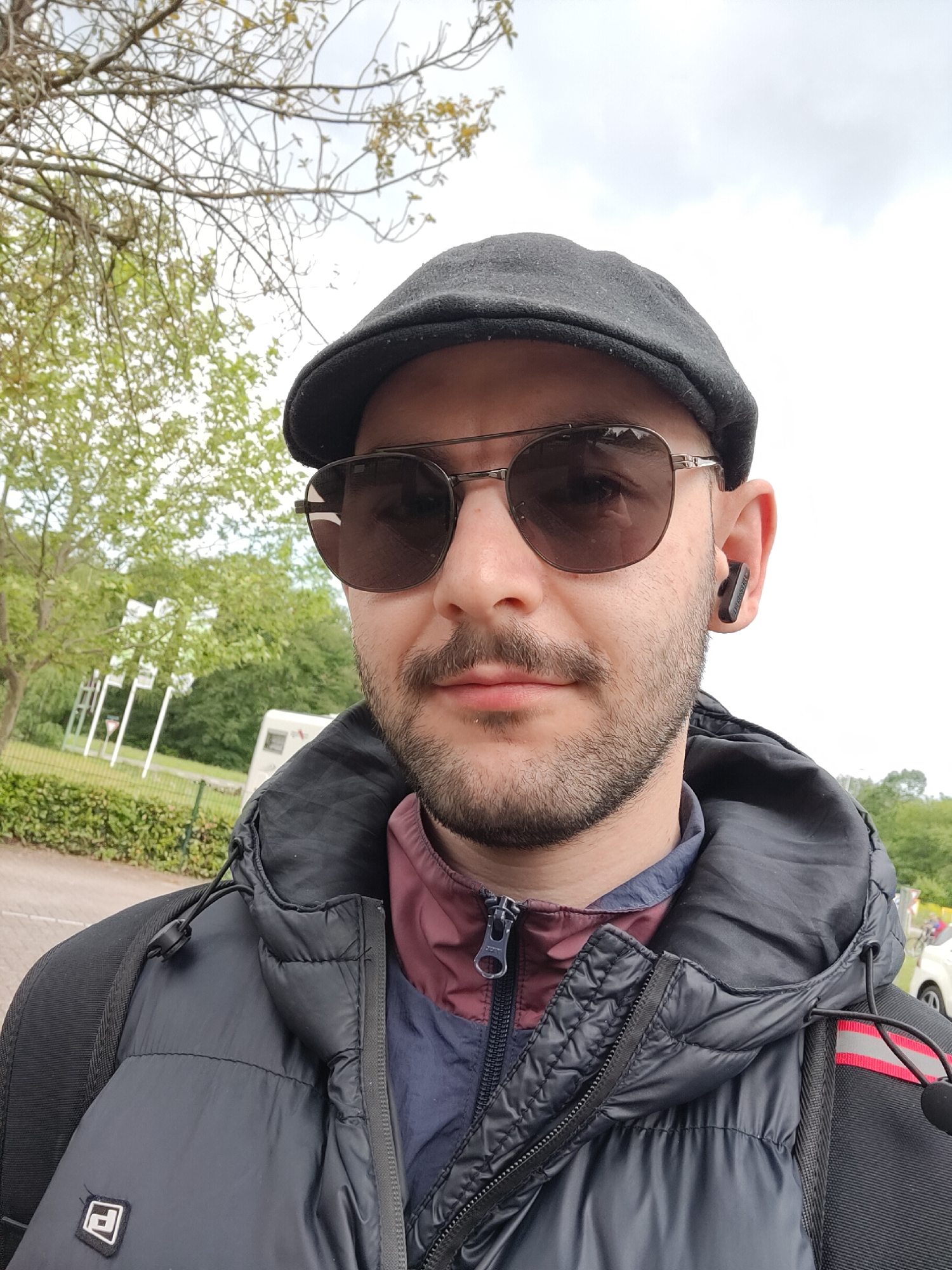 A close-up selfie depicts a man wearing a dark newsboy cap, aviator-style sunglasses, a dark puffer jacket over a layered shirt, and a black wireless earbud in his right ear. He has a mustache and a short beard.
The background reveals a partially overcast sky, trees, and a green grassy area with a hedge. In the distance, there is a white vehicle (possibly a campervan), and a signpost with multiple directional signs.
The location is likely in Oosterhout, Netherlands, based on the request, but there is no visual confirmation.
