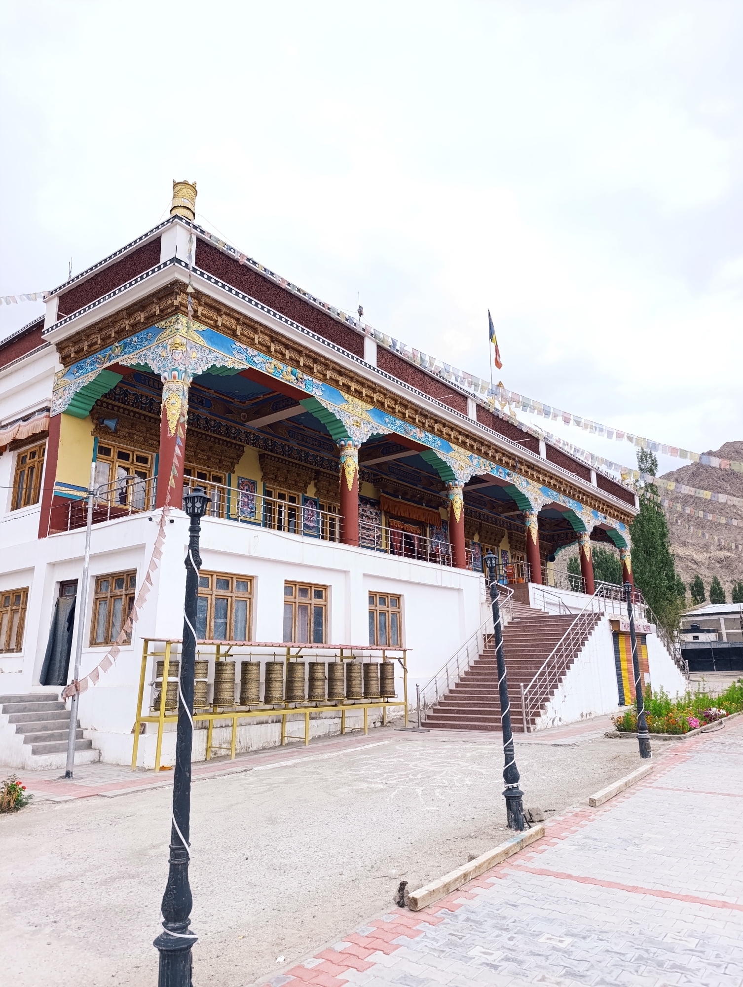 The image depicts the exterior of a Buddhist monastery in Leh, India. The building features traditional Tibetan architecture with white walls, ornate painted eaves, and a multi-tiered roof adorned with prayer flags. A prominent set of stairs leads up to the entrance. To the left of the stairs, a row of large, golden prayer wheels is visible, mounted on a yellow rack. Two black lampposts with decorative white bands stand in the foreground, lining a pathway paved with red and grey bricks. In the background, a mountainous landscape, also decorated with prayer flags, rises under a cloudy sky. The scene is devoid of people, creating a serene and contemplative atmosphere.
