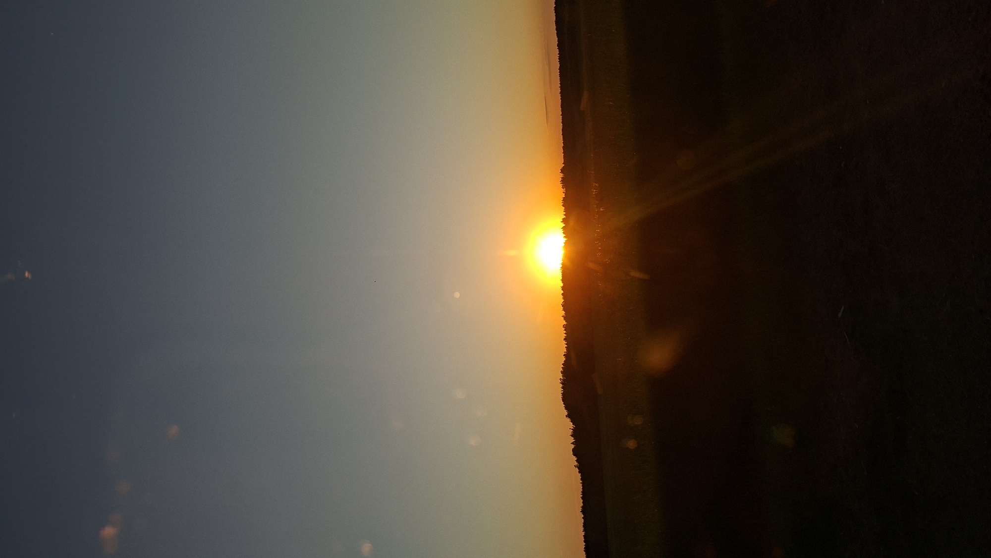 The image shows a sunset in Любанскі сельскі Савет, Belarus. The sun is setting on the horizon, with orange light. The foreground consists of dark land or foliage, while the sky transitions from dark blue to orange. There are some spots of light or lens flares scattered in the sky.