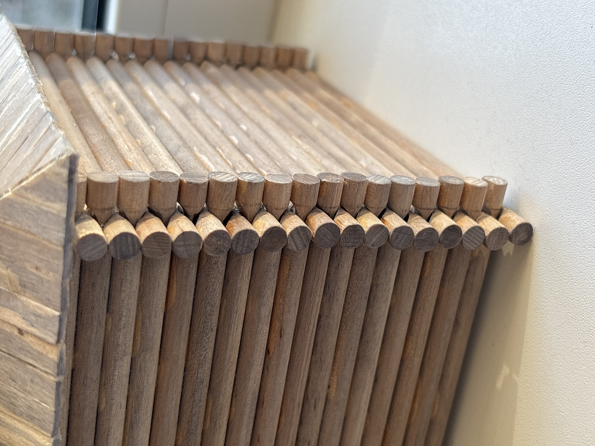 The image shows a close-up of a wooden structure, possibly a piece of furniture like a side table or storage unit, constructed from numerous dowels arranged in a parallel, repeating pattern. The dowels are light brown in color, and some are arranged horizontally and stacked, while others are placed vertically, creating the impression of walls. The dowels are joined together with wooden pins, and there is some slight wood grain.
The environment appears to be indoors, near a light-colored wall, implying that it is a modern design piece. The city and country could not be determined.