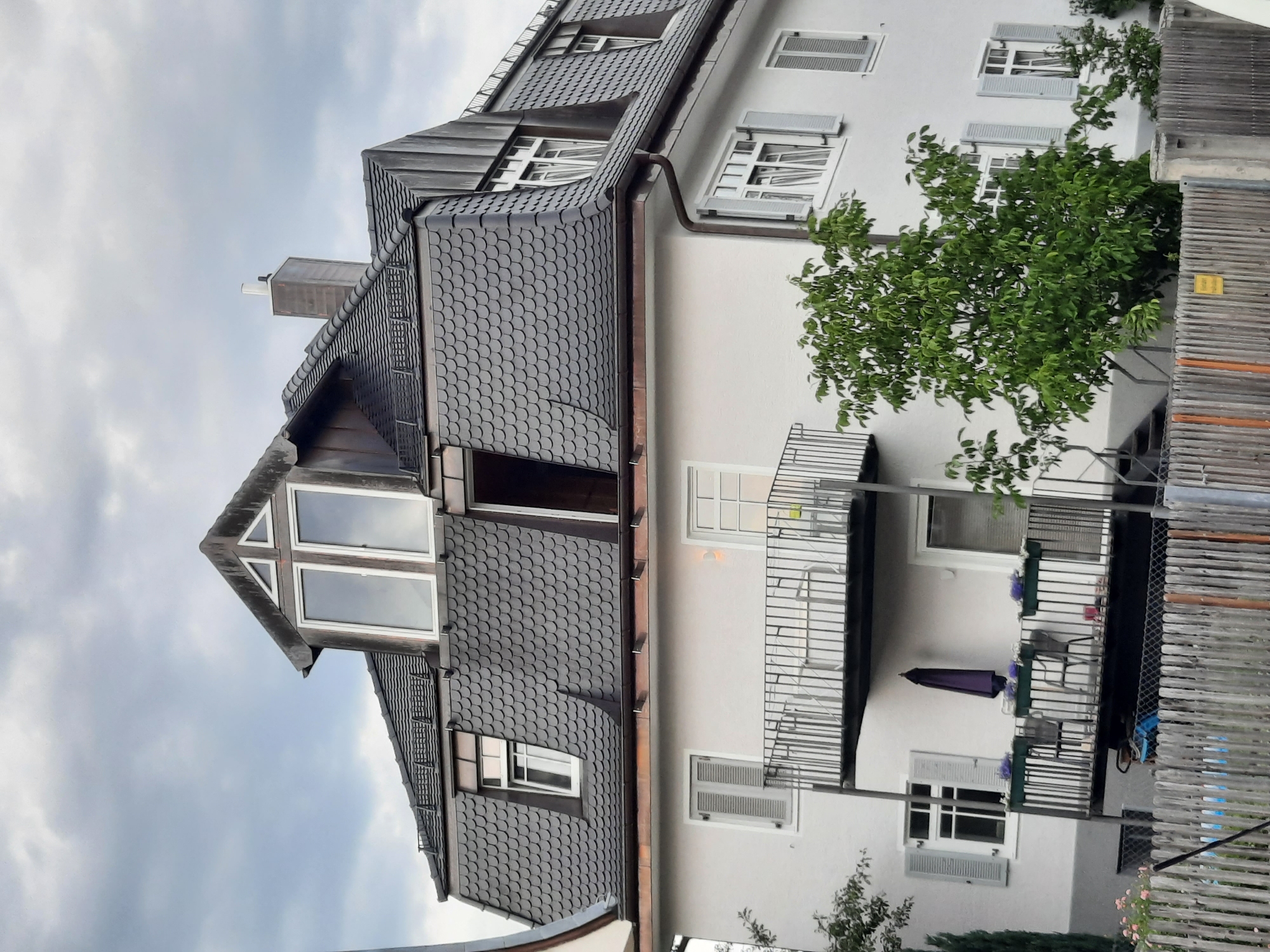 This image depicts a multi-story building with a dark, scale-patterned roof, characteristic of a mansard roof style, with dormer windows. The building's facade is painted white and features several windows with grey shutters and balconies. A prominent feature is a large tree with lush green foliage in front of the building. The sky above is overcast with grey clouds. A wooden fence runs along the bottom of the image. The scene is likely located in Rosenheim, Germany, given the architectural style and a yellow sign visible on the fence with German text. There are no people visible in the image.