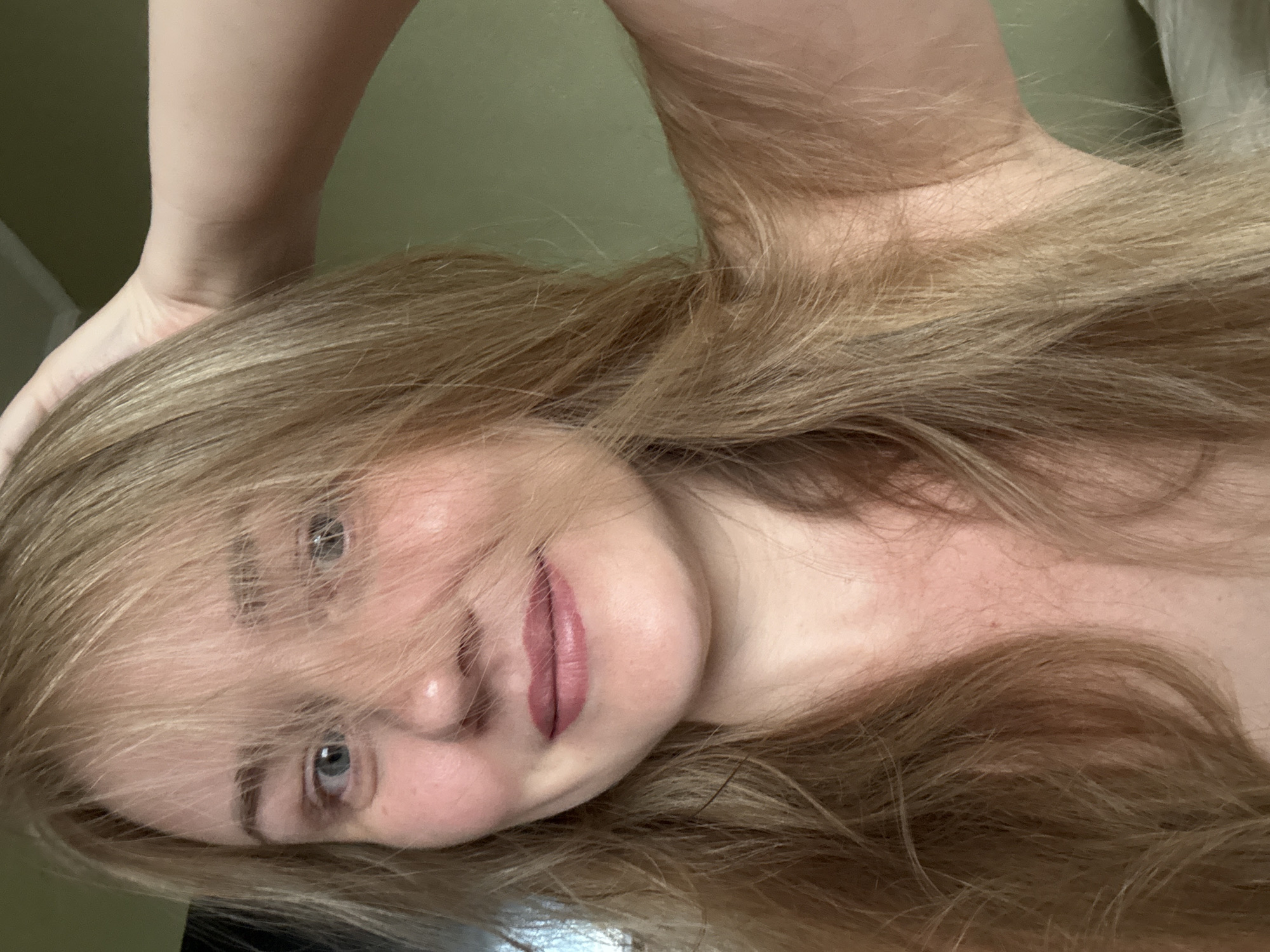 A woman with blonde hair is taking a selfie. She is looking directly at the camera with a slight smile. Her hair is partially covering her face. She is wearing a pink lipstick. She is raising her right hand to her head. The background is a plain green wall. This image was not taken in the city of Chaikovsky, Russia.