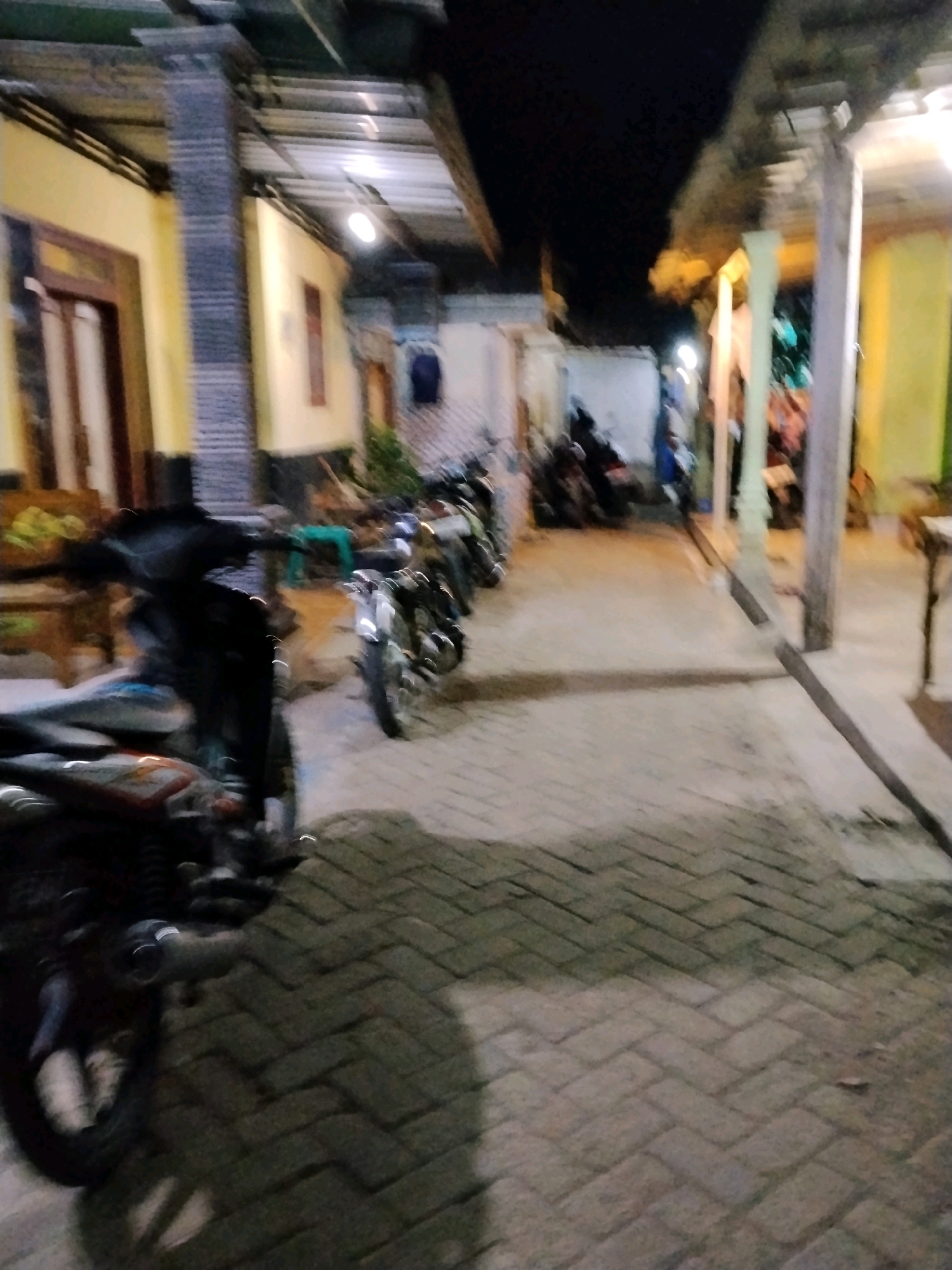 The image depicts an outdoor scene at night, possibly a residential alley or courtyard in Panggungan, Indonesia. The ground is paved with herringbone-patterned bricks. Several motorcycles are parked in a line along the right side of the alley. The buildings on either side of the alley have yellow walls and visible roof structures, with some lighting illuminating the area. The overall atmosphere is dim, suggesting it is late at night or early morning. No individuals are clearly visible in the frame, and no specific actions or interactions are evident.