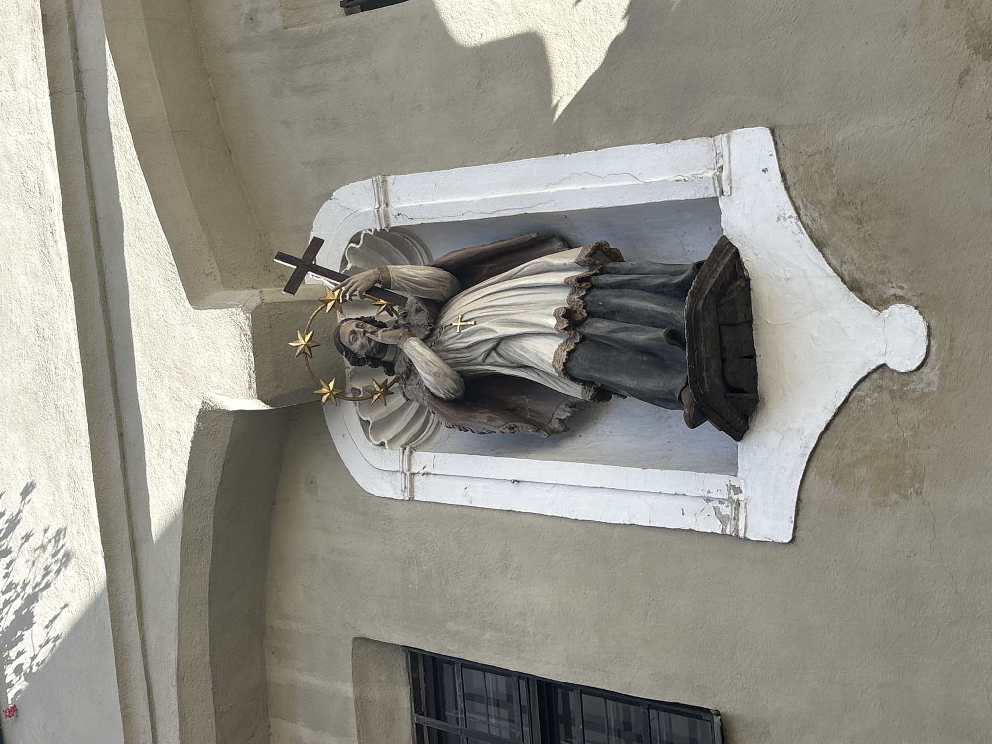 The image shows a statue of a saint, possibly John of Nepomuk, mounted on the exterior of a building in an unknown city. The saint is depicted in traditional garb, with a halo of stars, holding a cross. The statue is set within a decorative, white alcove attached to the building's wall. Below the statue, a window with black bars is visible. The building's exterior is painted a light neutral color, and the architectural style appears to be classical or baroque. The environment is urban, as evidenced by the presence of the building and window.