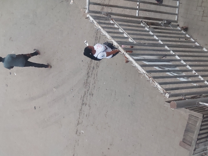 The scene, located in Agani, Nigeria, is an elevated view of a dusty, light-colored ground surface with scattered debris and faint tire tracks. Two individuals are present. In the upper frame, a male subject, viewed from the back, walks leftward, wearing a dark short-sleeved shirt, dark trousers, and light-colored shoes. In the middle frame, a female subject walks rightward, adjacent to a metal barrier. She has dark hair in a ponytail, wears a white short-sleeved t-shirt, dark lower garment, and light-colored sandals, carrying a dark shoulder bag. The metal barrier, with vertical bars and anti-climb spikes along its top, occupies the lower right. A wooden step structure is visible in the bottom-left corner.