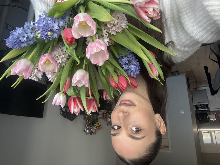 A female subject is positioned in the foreground of an indoor scene within a residential apartment in Sofia, Bulgaria. She has long, dark hair pulled back from her face and is wearing a light-colored, textured sweater. Her gaze is directed towards the camera. She holds a large bouquet of fresh flowers, prominently featuring pink and red tulips, interspersed with blue and light pink hyacinths. The background to the left reveals a modern kitchen area with white upper cabinets, a light countertop, and a built-in appliance. A dark chair is partially visible on light-colored wooden flooring. To the right, a dark flat-screen television is visible, along with a dark wooden console table displaying several decorative objects, including a metallic bull sculpture. The walls are a neutral light grey.
