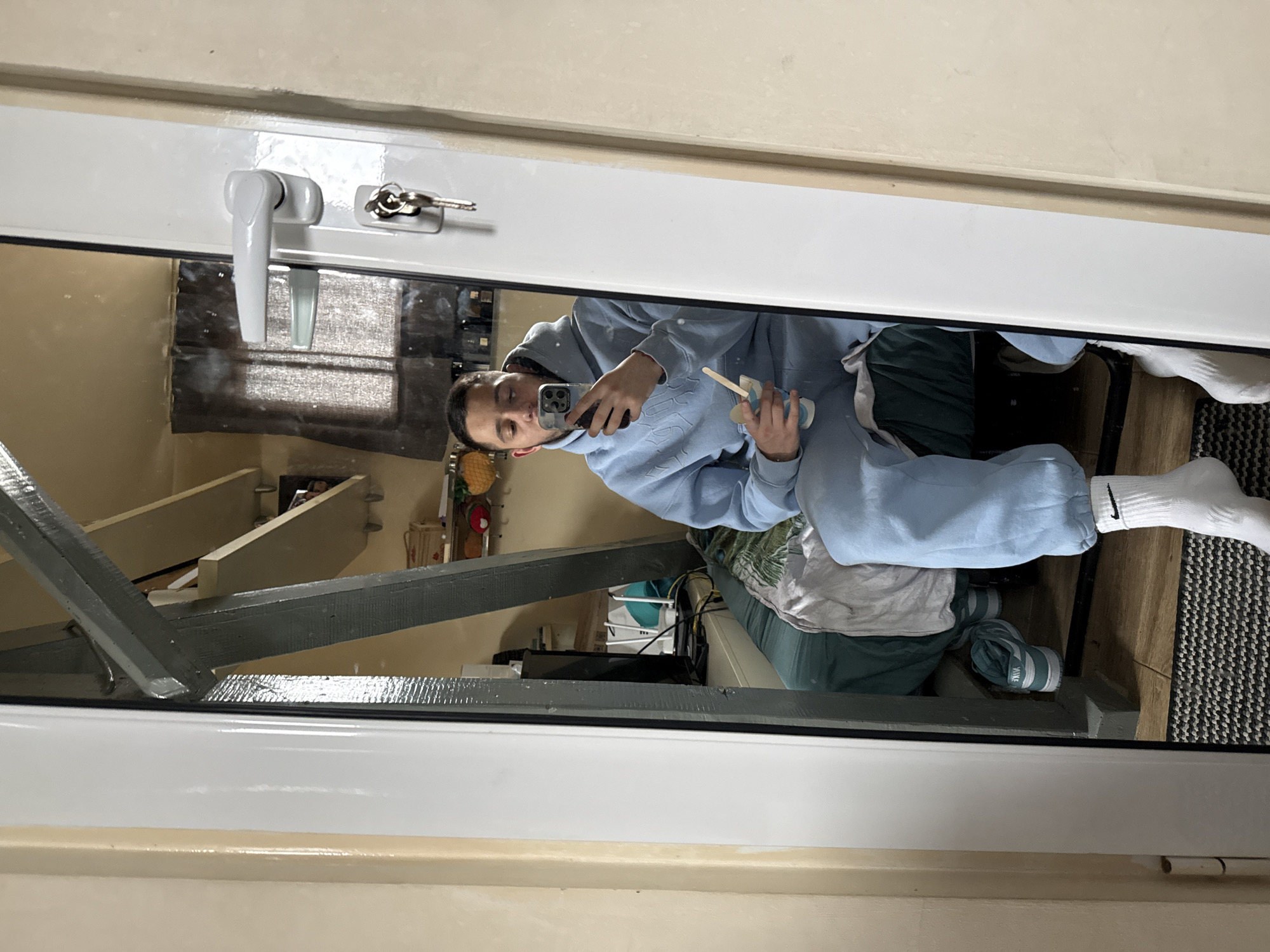 A young man is taking a selfie in a mirror. He is wearing a light blue sweatsuit and white socks. He is holding a phone in one hand and what appears to be a small cup and spoon in the other. The reflection shows a glimpse of a cluttered room with various items. The reflection of the room indicates a domestic setting. The mirror is likely situated on a window or door, indicated by the handle and locking mechanism visible at the top of the frame. Location information cannot be determined from this image.