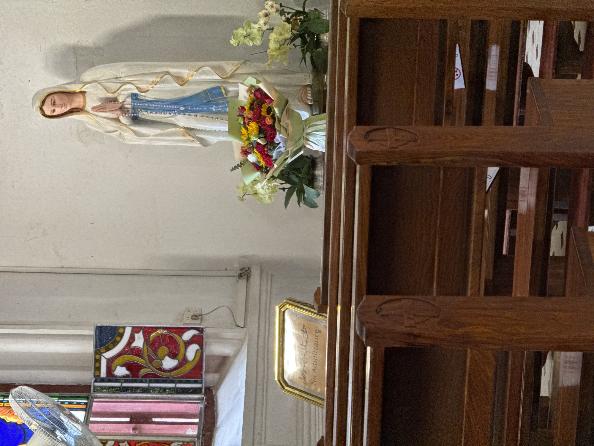 A photograph depicts the interior of a church in the Liwan District, China, oriented to a horizontal perspective. The left side of the scene features a statue of the Virgin Mary in a white and blue robe, adorned with a bouquet of colorful flowers placed at its base. The room's color scheme is primarily white, with a stained glass window visible underneath the statue. On the right side, church pews are arranged with wooden frames. A sign labeled "No Admittance" is affixed to one of the wooden structures. The scene appears to be well-lit.