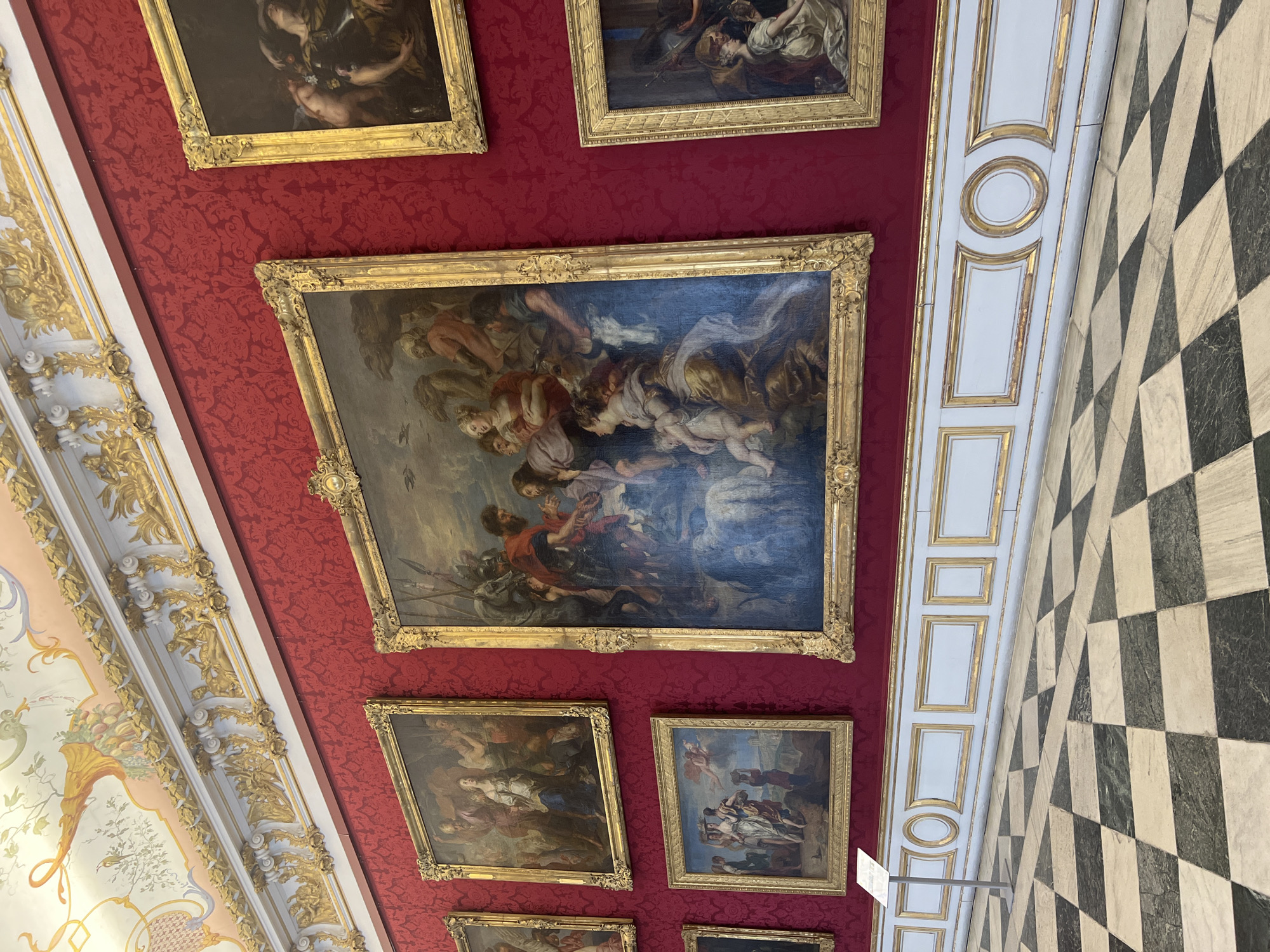 This image depicts a grand hall, likely within a palace or museum, in Oberschleißheim, Germany. The room features a richly decorated red damask wallpaper adorning the walls, accented by elaborate golden moldings and intricate ceiling frescoes. A series of large, ornate framed paintings hang on the wall, showcasing what appear to be historical or religious scenes with multiple figures. The paintings are displayed in a salon-style arrangement, with one particularly large canvas dominating the center.

In the foreground, a striking black and white checkered marble floor extends across the hall. A simple white display stand is positioned in front of one of the paintings. The lighting suggests an indoor setting, possibly illuminated by natural light filtering in from unseen sources, highlighting the opulent details of the architecture and artwork.