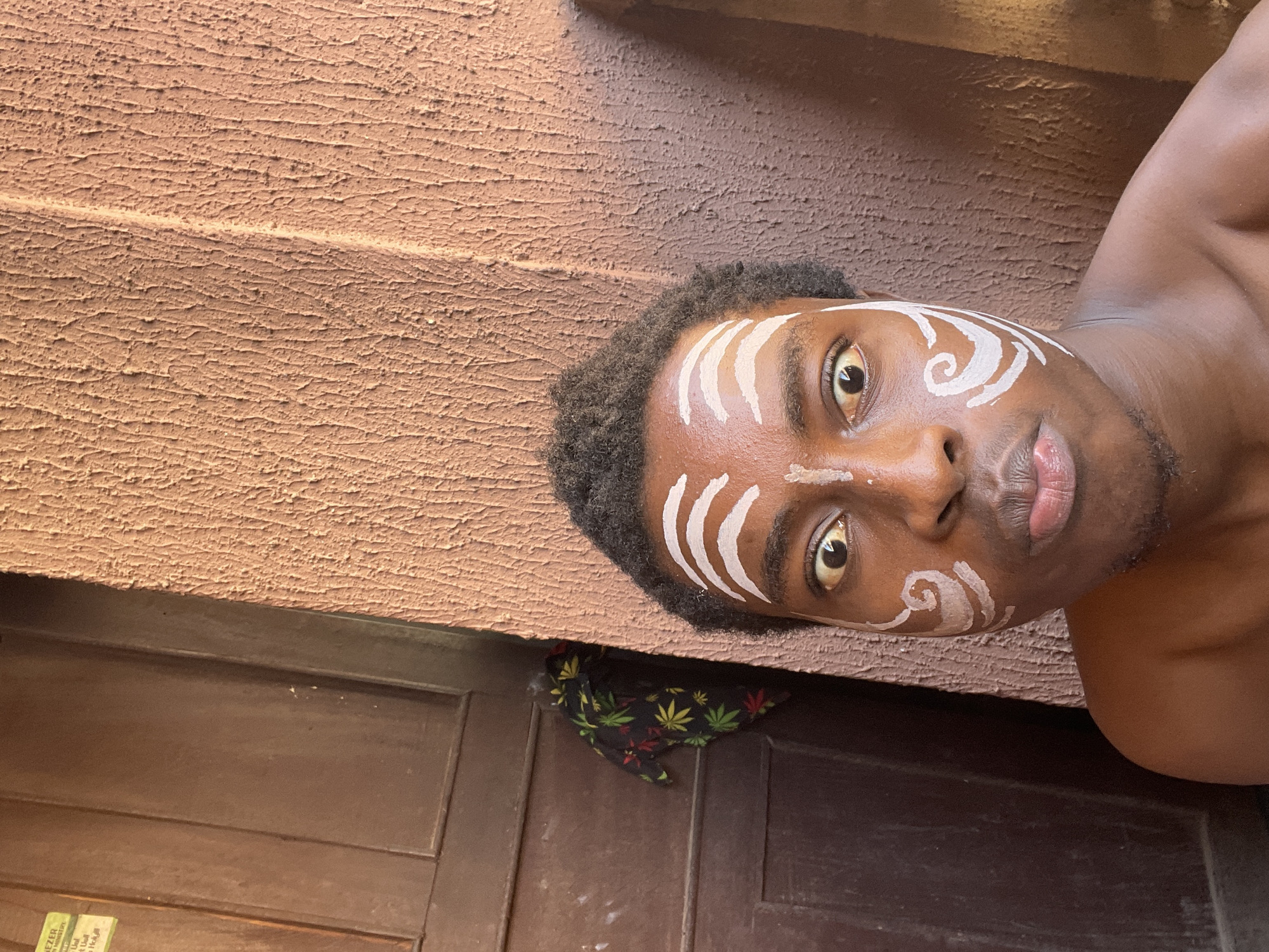 A close-up portrait, oriented sideways, features a young Black man. He is shirtless and has striking white paint designs on his face. Above his left eye (viewer's left), there are three distinct vertical white stripes. On the right side of his face, a prominent white spiral design extends from his temple towards his cheek. His short, dark, curly hair frames his forehead. The man has wide, dark eyes, looking directly forward, and his lips are slightly parted, revealing a neutral expression.
The background primarily consists of a rough, light reddish-brown textured wall, occupying the upper and left parts of the frame. Along the bottom edge of the image is a dark brown wooden door with visible paneling. A small piece of black fabric, patterned with green, red, and yellow marijuana leaves, is visible peeking out from the gap between the wall and the door. The lighting suggests natural daylight. On a small, partially visible white label or sticker near the bottom left, faint text like "EZER", "services", "Usul", and "tea" can be discerned. The setting, located in Nkpor, Nigeria, suggests a cultural or artistic context for the facial paint.