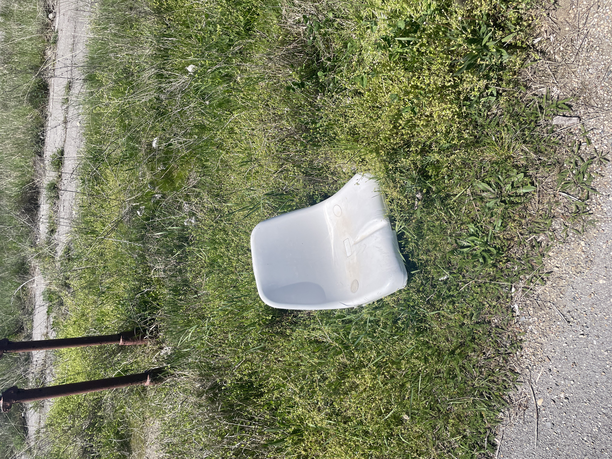 A discarded white plastic chair sits in green grass next to a concrete curb. Two rusted metal pipes lie on the ground near the grass.
I cannot identify the location as Sofia, Bulgaria, as there are no landmarks indicating its location.