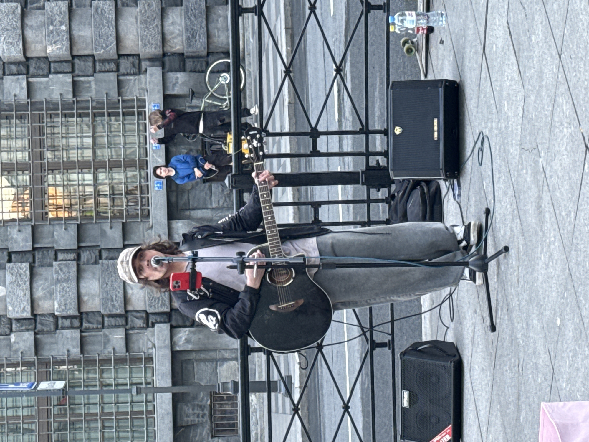 The image shows a street musician performing in front of a building with a textured stone facade. The musician, wearing a cap and a dark jacket over a light-colored shirt and grey pants, is playing an acoustic guitar and singing into a microphone. A red smartphone is attached to the microphone stand, possibly for lyrics or recording. In the background, two other individuals are visible near a bicycle. The scene is set on a paved urban street with a metal railing separating the performance area from the road. Two black speaker cabinets are positioned on either side of the musician. The overall setting suggests a public space in a city, likely a pedestrian area where street performers are common. The architectural style of the building, with its repeating rectangular stone blocks and barred windows, could indicate a historical district, potentially in Saint Petersburg, Russia, known for its grand architecture.
