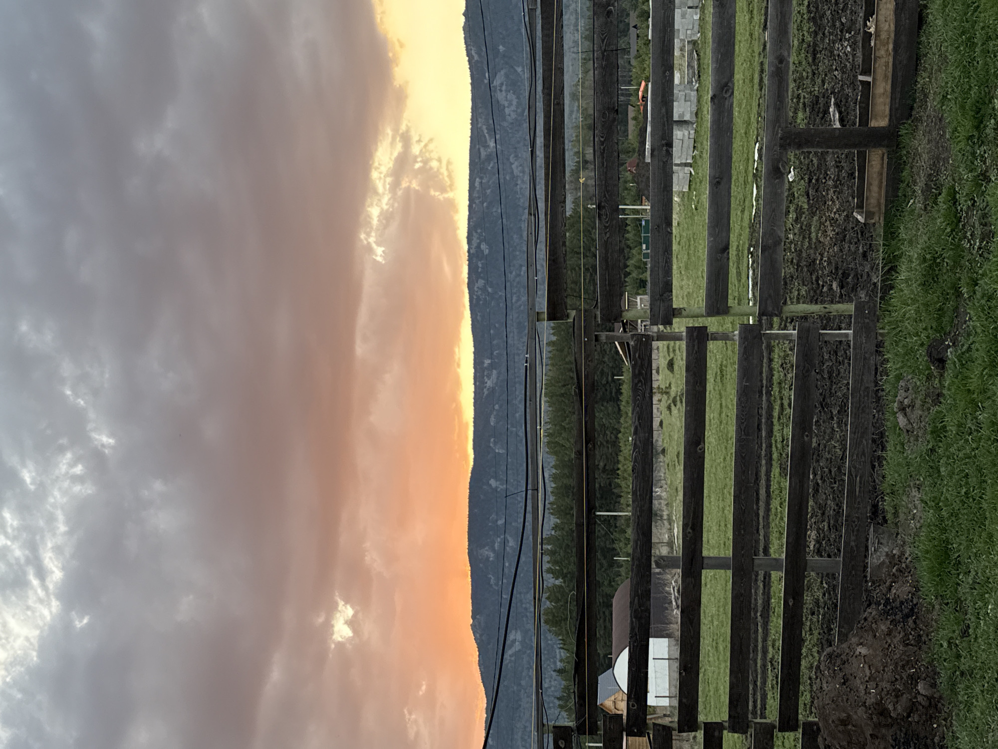 The image captures a rural scene in Tyulyuk, Russia, likely at dusk or dawn. A wooden fence runs vertically on the right side of the frame, with sections of open field and muddy ground behind it. Beyond the fence, there's a treeline and a mountain range visible in the distance. Above the mountains, the sky is filled with clouds, colored in shades of grey and orange, suggesting either a sunrise or sunset. Power lines stretch horizontally across the frame.