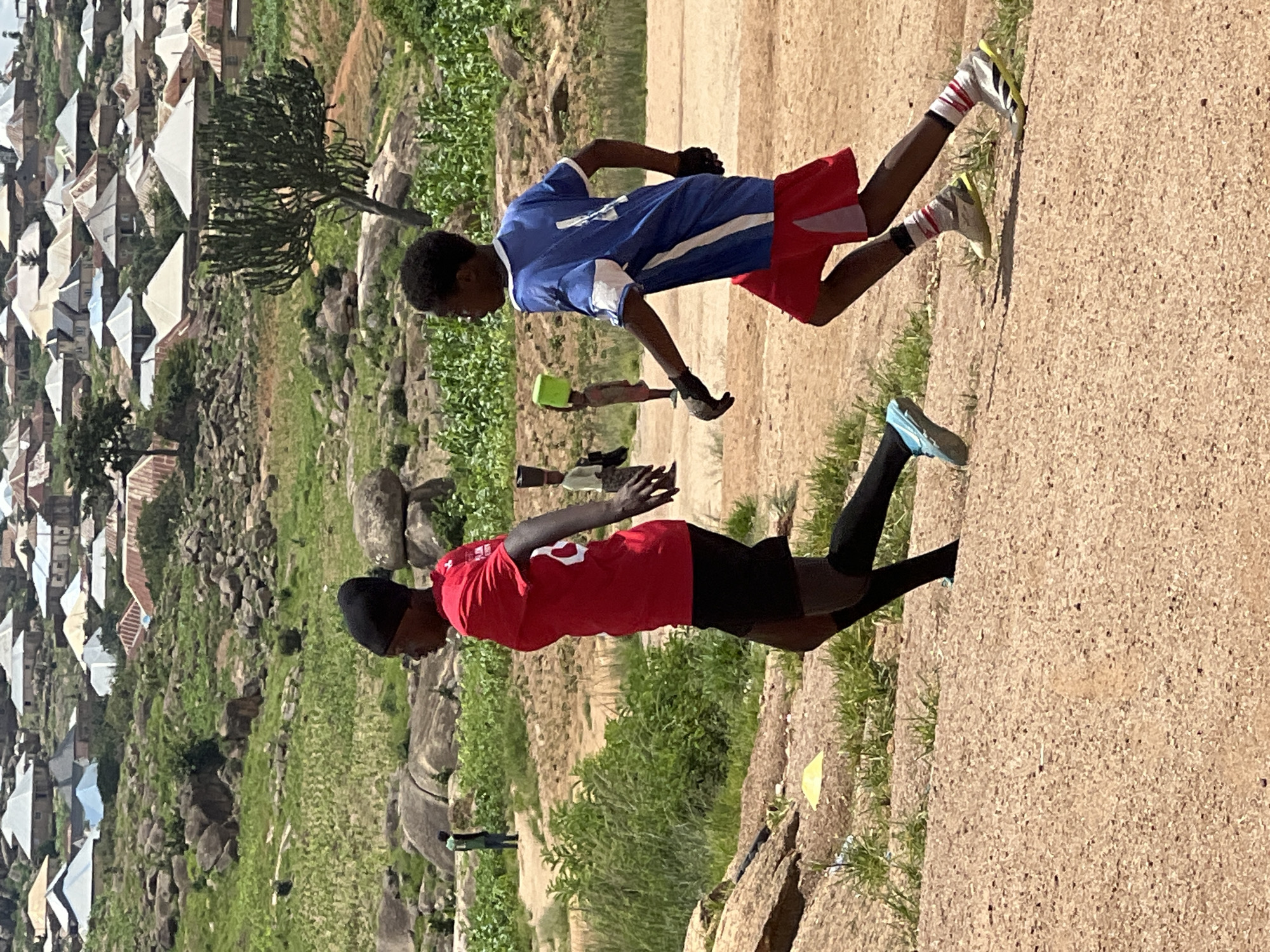 This image depicts two young individuals, likely playing soccer, on a dusty, uneven terrain in a rural or semi-rural setting with a sprawling settlement in the background. The person in the foreground on the left wears a red t-shirt with white markings, black shorts, and black socks. They have a black head covering and are in motion, appearing to run. The second person, on the right, is wearing a blue and white soccer jersey, red shorts, and white socks with red stripes. They are also running, with black gloves on their hands. The environment is characterized by scattered rocks and sparse vegetation, with a noticeable cluster of buildings in the distance, suggesting a community or village. The settlement in the background features numerous houses with corrugated metal roofs of varying colors, indicating a densely populated area. The overall impression is one of outdoor activity within a community that appears to be situated in a hilly or rocky landscape. The scene is likely located in the city of Jos, Nigeria, known for its hilly topography and diverse settlements.