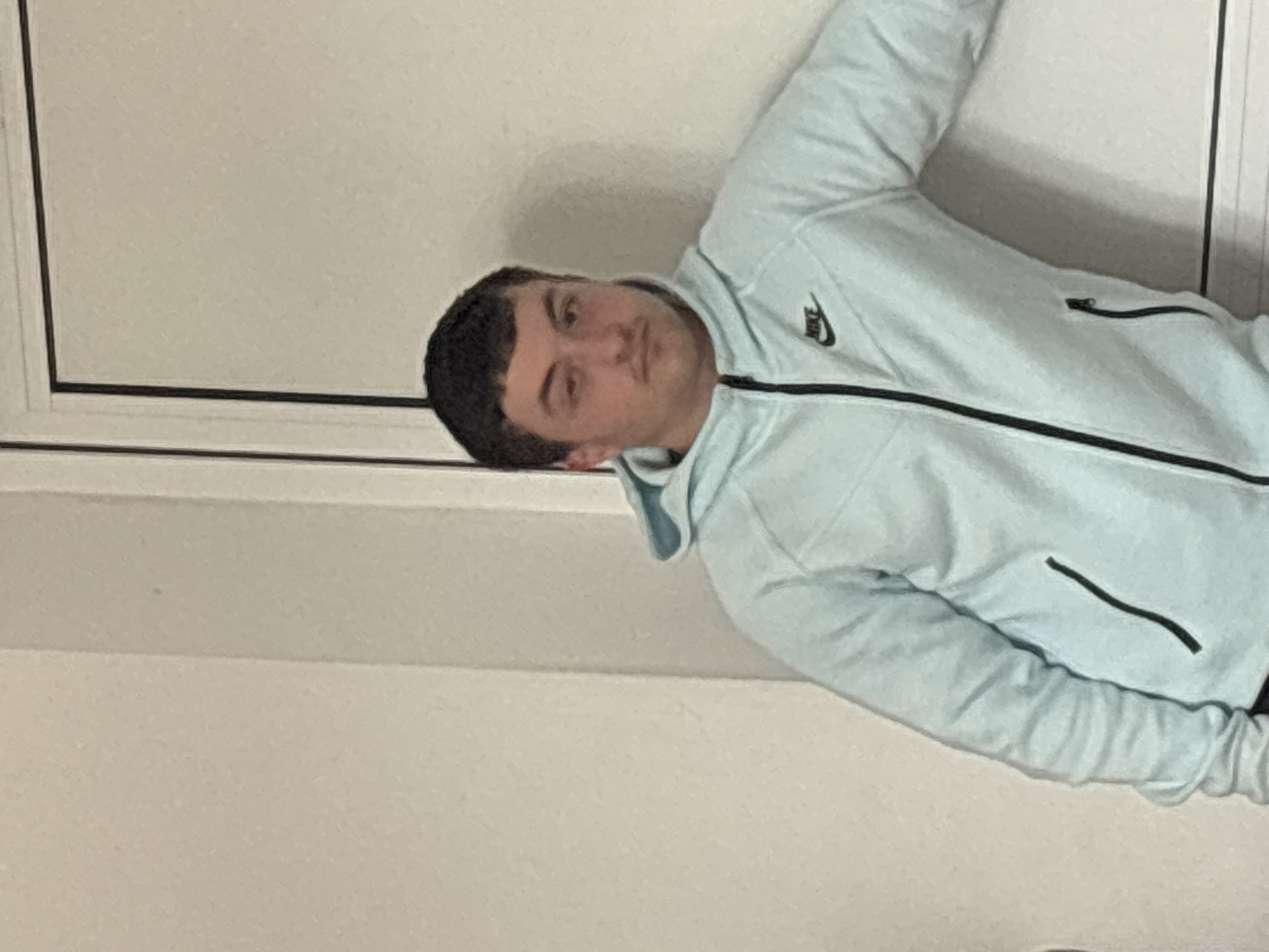 The image features a young man indoors in Zlatograd, Bulgaria, with his head tilted to the left. He is looking directly at the camera with a neutral expression. He has short, dark hair and appears to be leaning or posing against a light-colored wall. He is wearing a light blue Nike hooded jacket with black trim along the main zipper and pockets. The iconic Nike swoosh logo is visible in black on the left chest of the jacket. One of his arms is extended upwards, partially out of the frame. The background consists of plain, light-colored walls, with a distinct black line running vertically and horizontally on the left side, suggesting a door frame or wall trim. The lighting is consistent with an indoor environment, and no specific time of day is discernible.