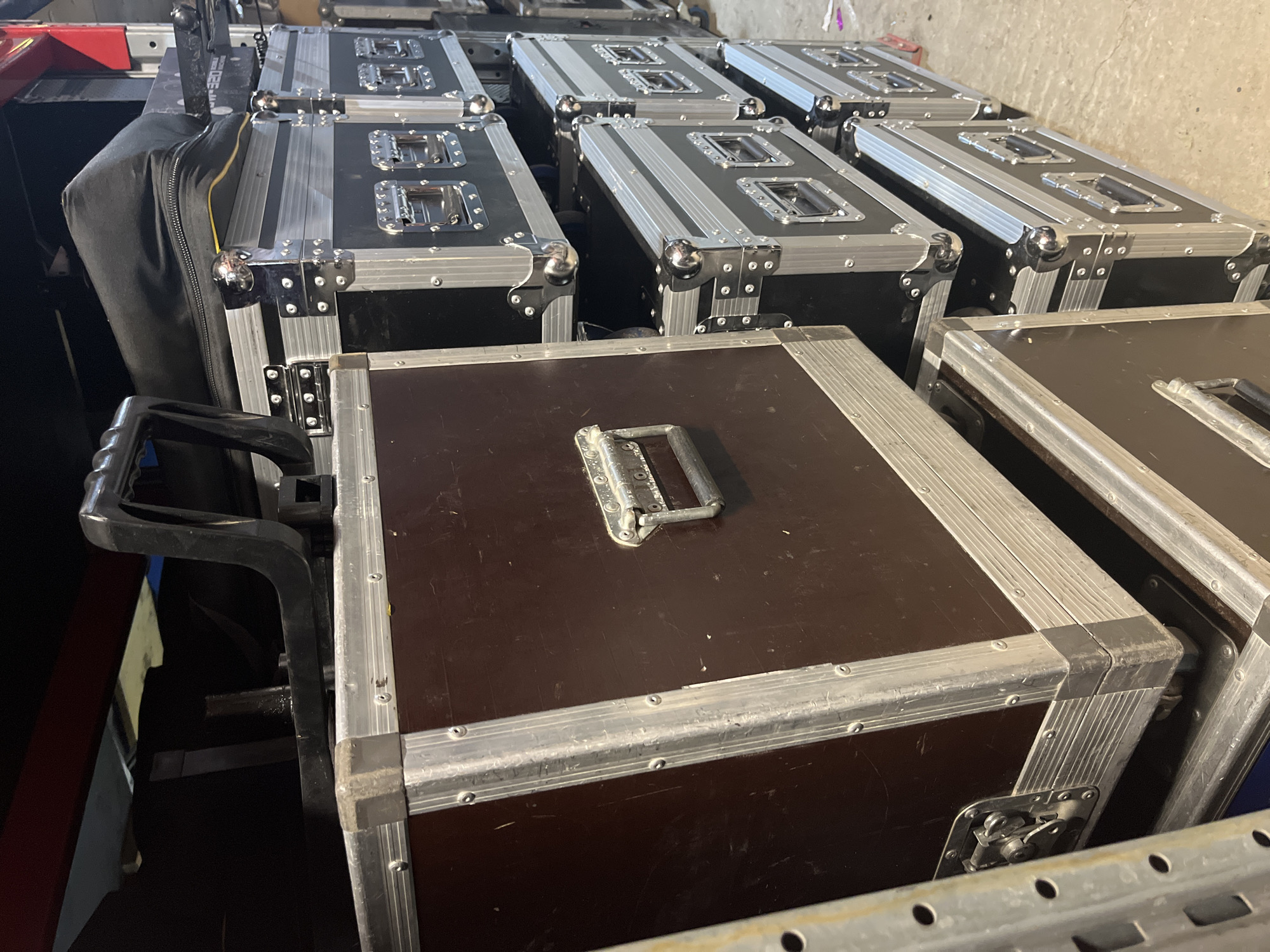 The image shows a storage area with multiple large, dark brown and black flight cases stacked on shelves. The flight cases have silver metal trim and latches. One large flight case is in the foreground on the left, and several smaller flight cases are stacked to its right, extending into the background. Some of the flight cases have handles visible. The lighting is dim and appears to be artificial. The environment seems to be a warehouse or storage facility.