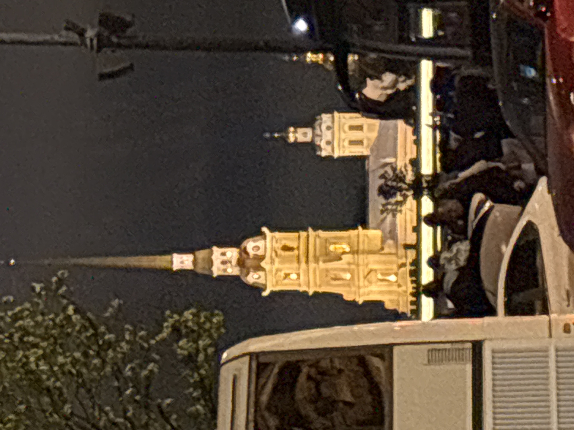 The image depicts the Peter and Paul Cathedral in Saint Petersburg, Russia, viewed at night. The illuminated spire and tower of the cathedral are prominent against the dark sky. Part of a vehicle, possibly a bus or large car, is visible in the foreground. To the left, foliage from a tree is partially visible. The shot is taken from a slightly elevated perspective, possibly from inside or atop another vehicle.
