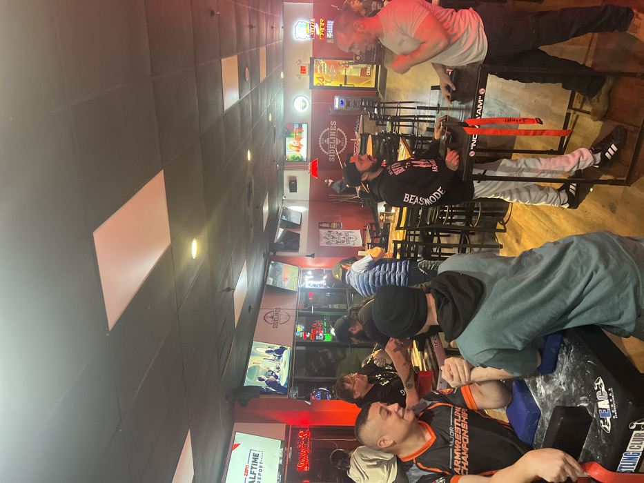 Inside a brightly lit establishment, possibly a sports bar, several individuals are engaged in arm wrestling matches. The scene includes multiple arm wrestling tables set up, with participants facing off. Televisions displaying "SIDELINES" and "ESPN HALFTIME REPORT" are visible in the background. Signage references "MAJOR ARMRESTLING CHAMPIONSHIP," indicating an organized event. Participants wear casual attire, and the atmosphere appears lively and competitive.