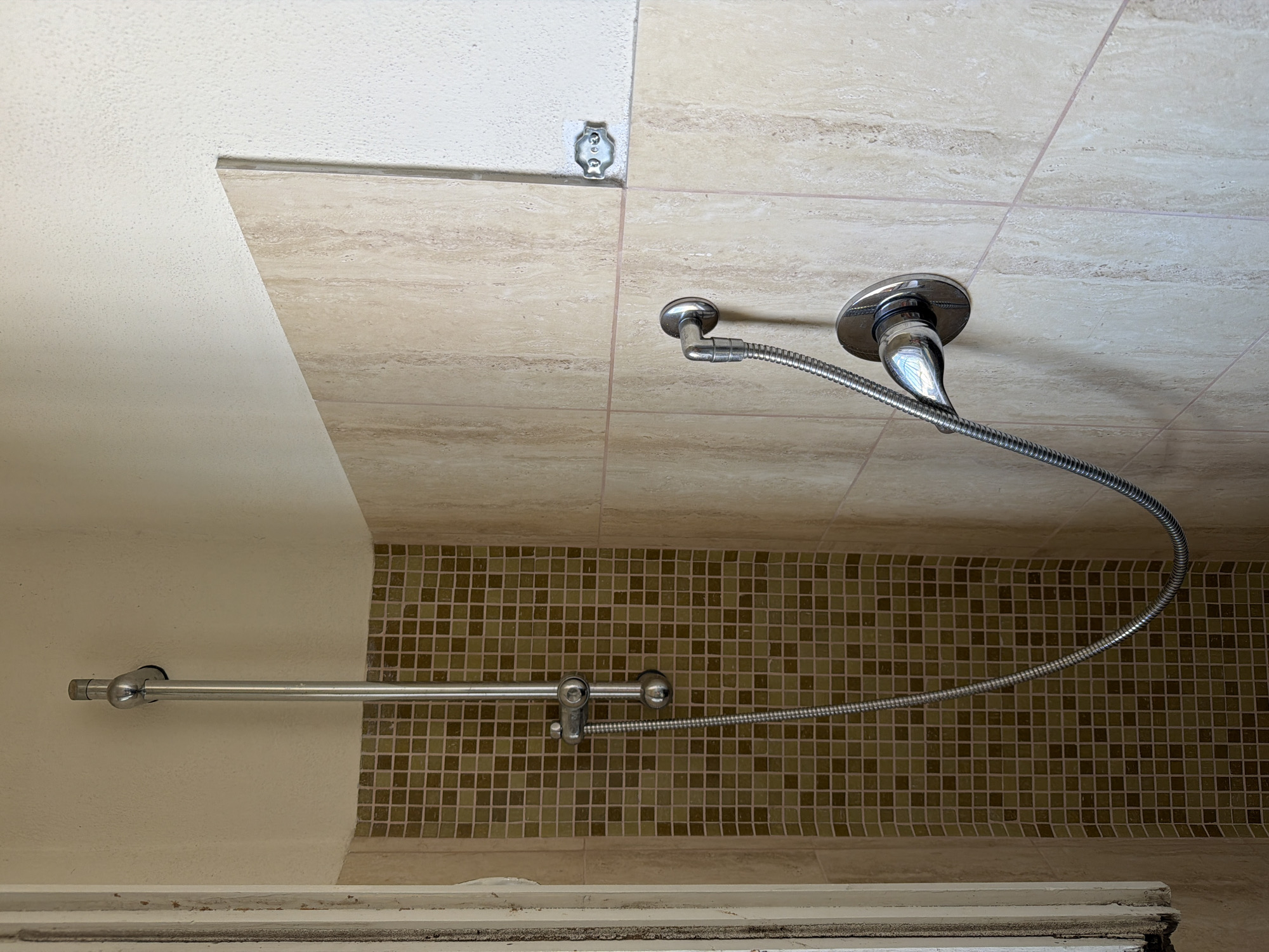 The image shows a close-up view of a shower area in a bathroom. A chrome showerhead is mounted on a vertical chrome pipe, which is attached to the wall. A flexible chrome hose connects the showerhead to a control handle on the wall. The walls are partially tiled with large, light-colored travertine tiles and partially with small, dark brown mosaic tiles. The grout lines of the tiles are visible. The scene is well-lit, suggesting an indoor environment. There are no people or animals visible in the image. The overall impression is of a functional, though perhaps dated, shower installation. The image does not provide specific visual cues to definitively place its location within the city of Olbia, Italy, beyond the fact that such a bathroom could exist in any urban or residential setting.