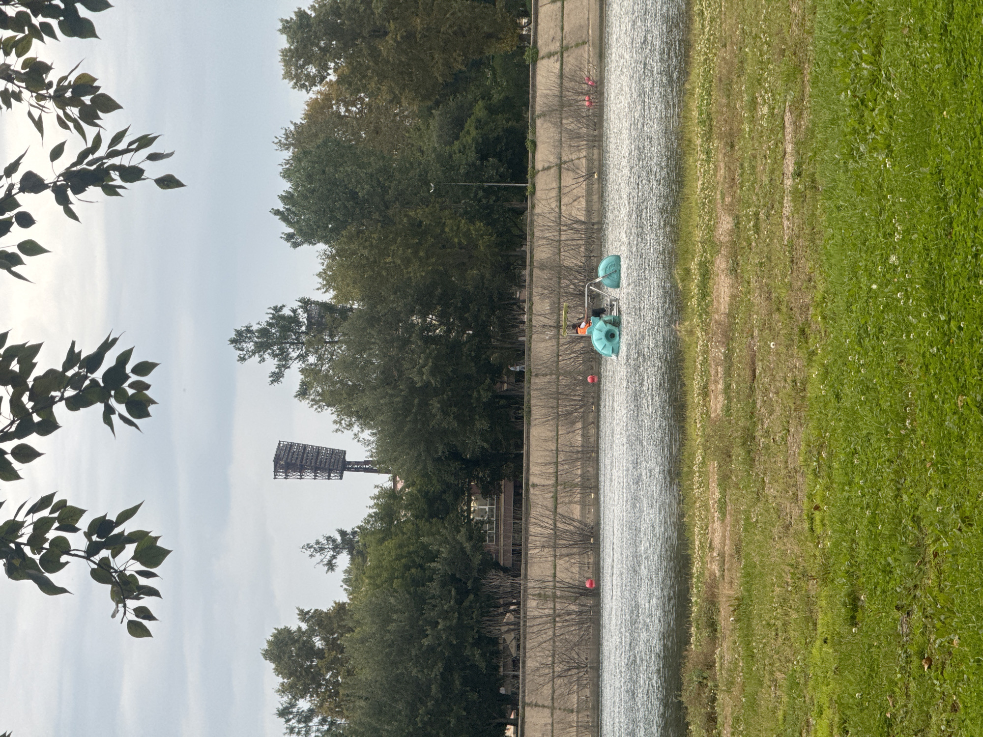 The image depicts a water body with a person operating a pedal boat. The boat is green with two pontoons and has a person in an orange vest seated at the controls. The water is choppy and reflects the overcast sky. In the background, a concrete embankment lined with leafless bushes separates the water from a grassy area with trees. Beyond the trees, a tall stadium light structure is visible, along with buildings and more trees. The foreground is a grassy field with scattered white flowers. The scene is likely a park or recreational area near a river or lake. The stadium light suggests a proximity to a sports facility. Without specific landmarks, pinpointing the exact location in Irkutsk, Russia is not possible from the visual information alone, but it appears to be a waterfront area within the city.