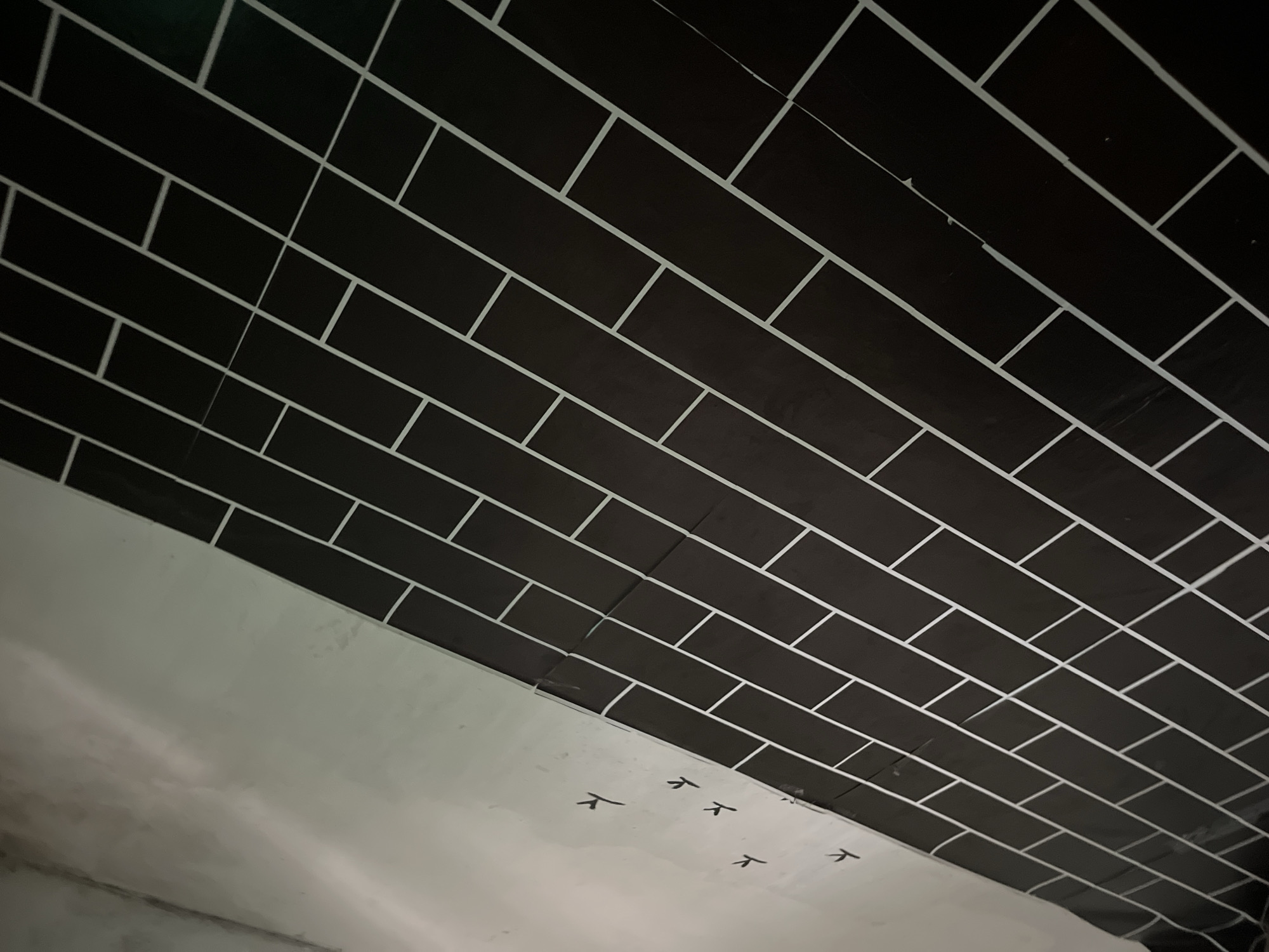 This is a close-up, angled shot of a wall with black brick-like tiling with white grout lines. The wall appears to be in an indoor setting, possibly under construction or renovation, as indicated by the unpainted section on the left and the faint black markings resembling bird footprints. The lighting is dim, casting shadows that highlight the texture of the wall. The image's perspective suggests it was taken from a low angle, looking upwards. The specific location of Krucil, Indonesia, is not discernible from the visual information in the image.