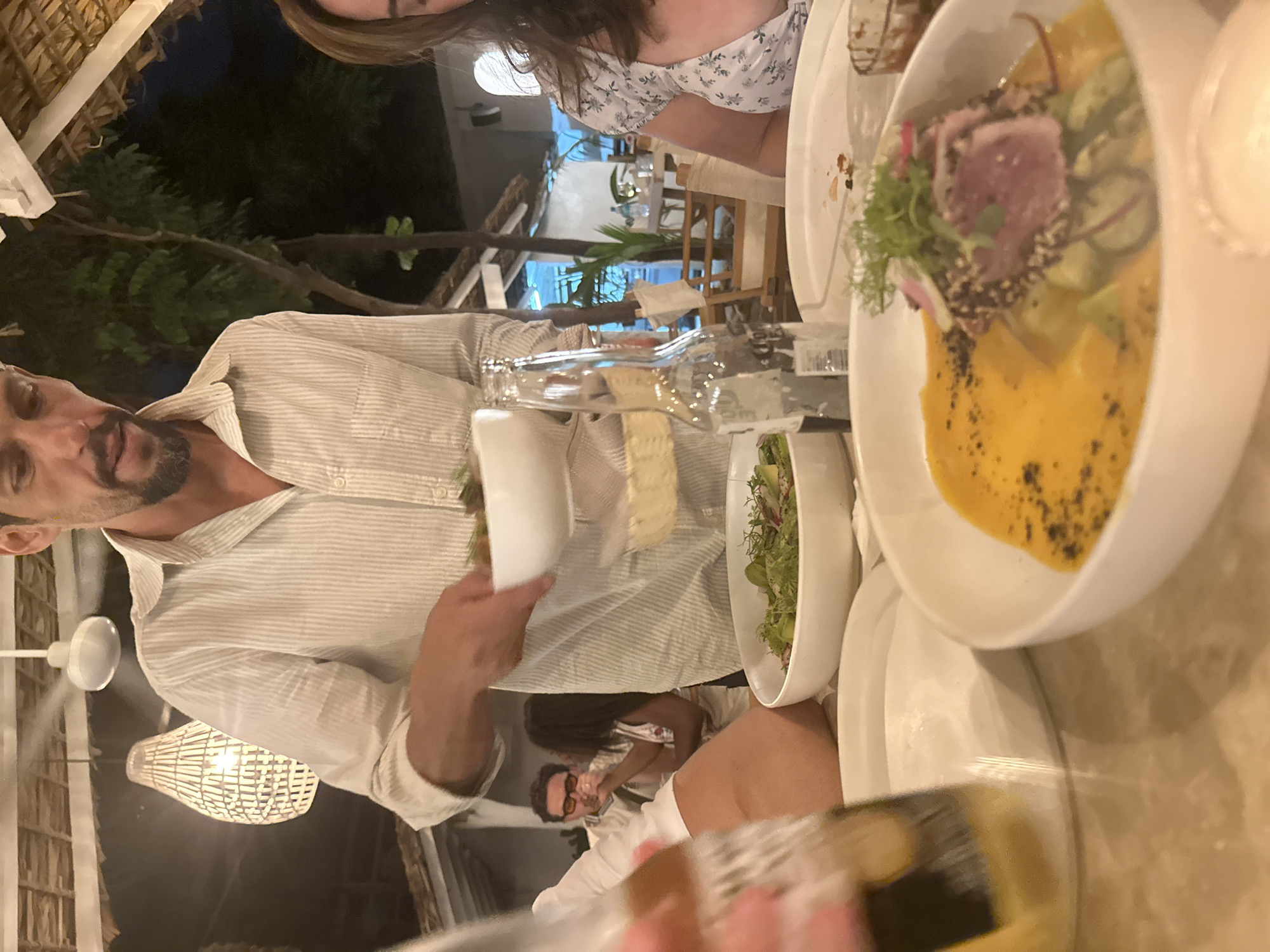 A group of people are seated at an outdoor dining table at night. The location appears to be a restaurant in Brisas de Zicatela, Mexico, evidenced by the beach setting and architecture. A man with a beard is wearing a striped shirt, holding a plate, and seemingly engaged in a conversation with other people at the table. A woman in a floral-patterned dress is partially visible near the top of the frame. Various dishes, including a salad and a tuna dish with mango and avocado, along with water bottles, are arranged on the table. Another person is visible wearing a mask near the bottom of the frame. The lighting includes a combination of natural light from the surroundings and artificial lighting from a ceiling fan and decorative lamps.