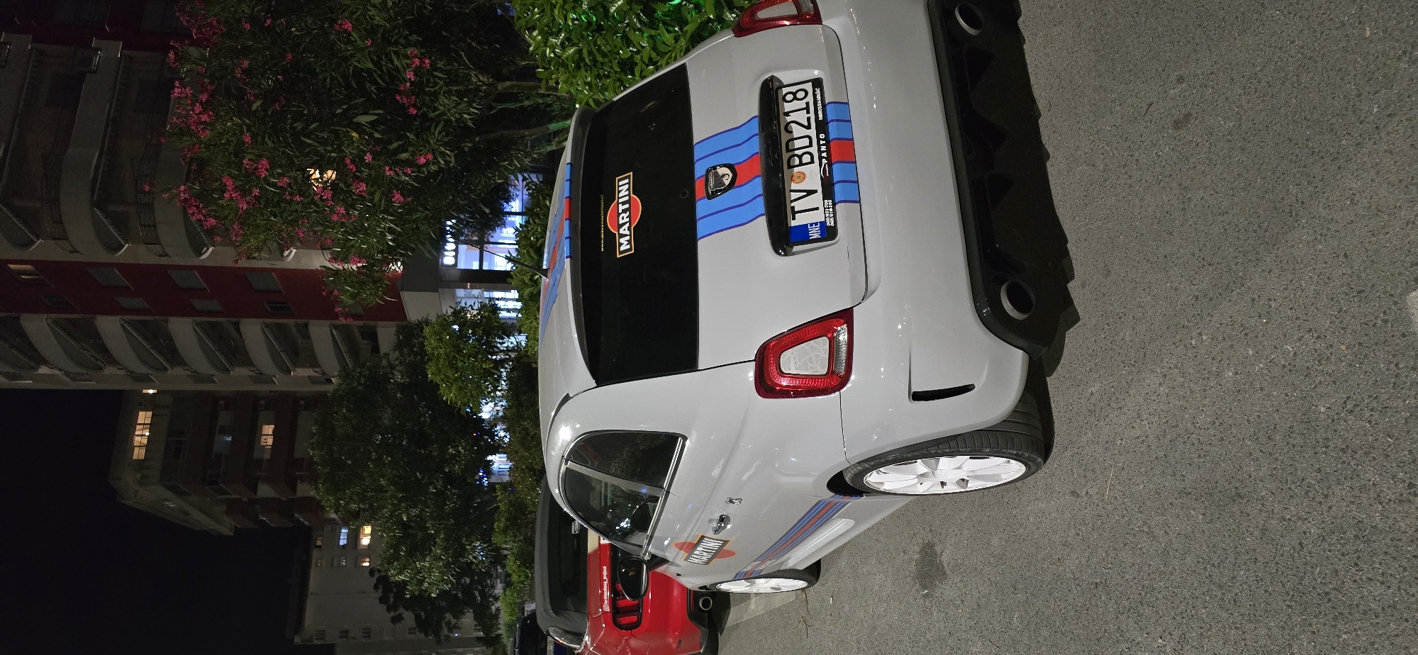 The image shows the rear of a white Fiat 500 Abarth parked on the street in Bar, Montenegro. The car has Martini racing stripes and decals. The license plate reads "TV BD218." Behind the Fiat, the front of a red car is visible. In the background are trees, a building, and streetlights. The scene appears to be at night.