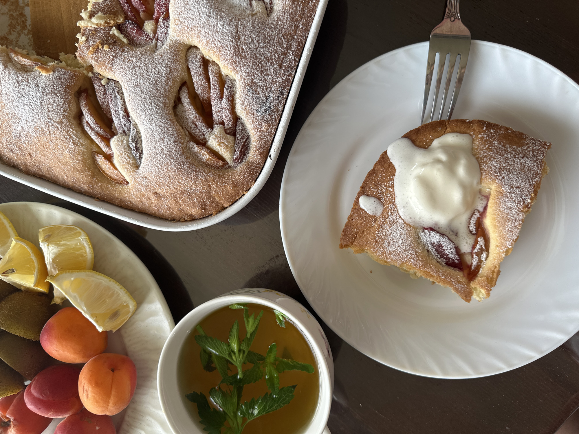 The image depicts a table setting, suggestive of a light meal or dessert, with a focus on baked goods and fresh produce.
A rectangular baking dish containing a partially eaten cake, dusted with powdered sugar and featuring plum slices, is situated at the upper-left corner of the frame. Adjacent to it is a white plate containing a slice of the same cake, topped with a dollop of cream and a fork.
To the left, a white plate is arranged with kiwi fruit, lemon wedges, and several apricots. A cup of tea, garnished with fresh mint leaves, is positioned between the plate of fruit and the slice of cake.
The table has a dark, possibly wooden, surface. The scene is captured with a direct overhead perspective, allowing a clear view of the arrangement.
The image offers no explicit indication of the location in the city of Chaikovsky, Russia.
