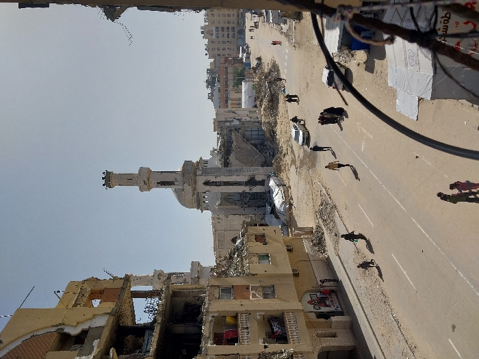 The image depicts a street scene in Khan Yunis, Palestinian Territory, characterized by significant structural damage to buildings under a clear sky.
In the foreground, a dark, partially blurred wire or cable extends from the lower right corner, partially obscuring a makeshift shelter covered with white tarpaulin.
To the left, a multi-story building exhibits extensive damage, with large sections of its facade crumbling, exposing internal floors and structural elements. Multiple impact holes are visible on its exterior. Laundry hangs from balconies on several floors. The ground floor features a storefront with visible graphics. Debris and rubble are scattered along the building's base and the unpaved roadside.
The central feature is a paved two-lane road with faded white lane markings. Several individuals are present on and alongside the road: a group of approximately three to four people, including at least one child, stands in the lower left; a person in dark clothing walks away from the camera followed by a smaller figure; a person in a light-colored top walks towards the background; and a cluster of several people, mostly in dark attire, are gathered on the right side of the road. Further down the street, additional individuals are walking. Two white sedans are visible; one parked on the right side of the road and another partially obscured behind the cluster of people. Another dark-colored vehicle is also present. The areas immediately adjacent to the road are unpaved, dusty, and covered with substantial amounts of rubble and debris.
In the mid-ground and background, a large, light-colored mosque with a tall minaret and a dome is prominent. The mosque itself shows considerable damage, particularly on its left side, where large sections of its structure are missing or collapsed. The minaret also displays damage towards its top. Debris surrounds the mosque's base. Beyond the mosque, other multi-story buildings, some appearing residential, are visible, many also exhibiting signs of damage or partial collapse. The distant background is hazy.