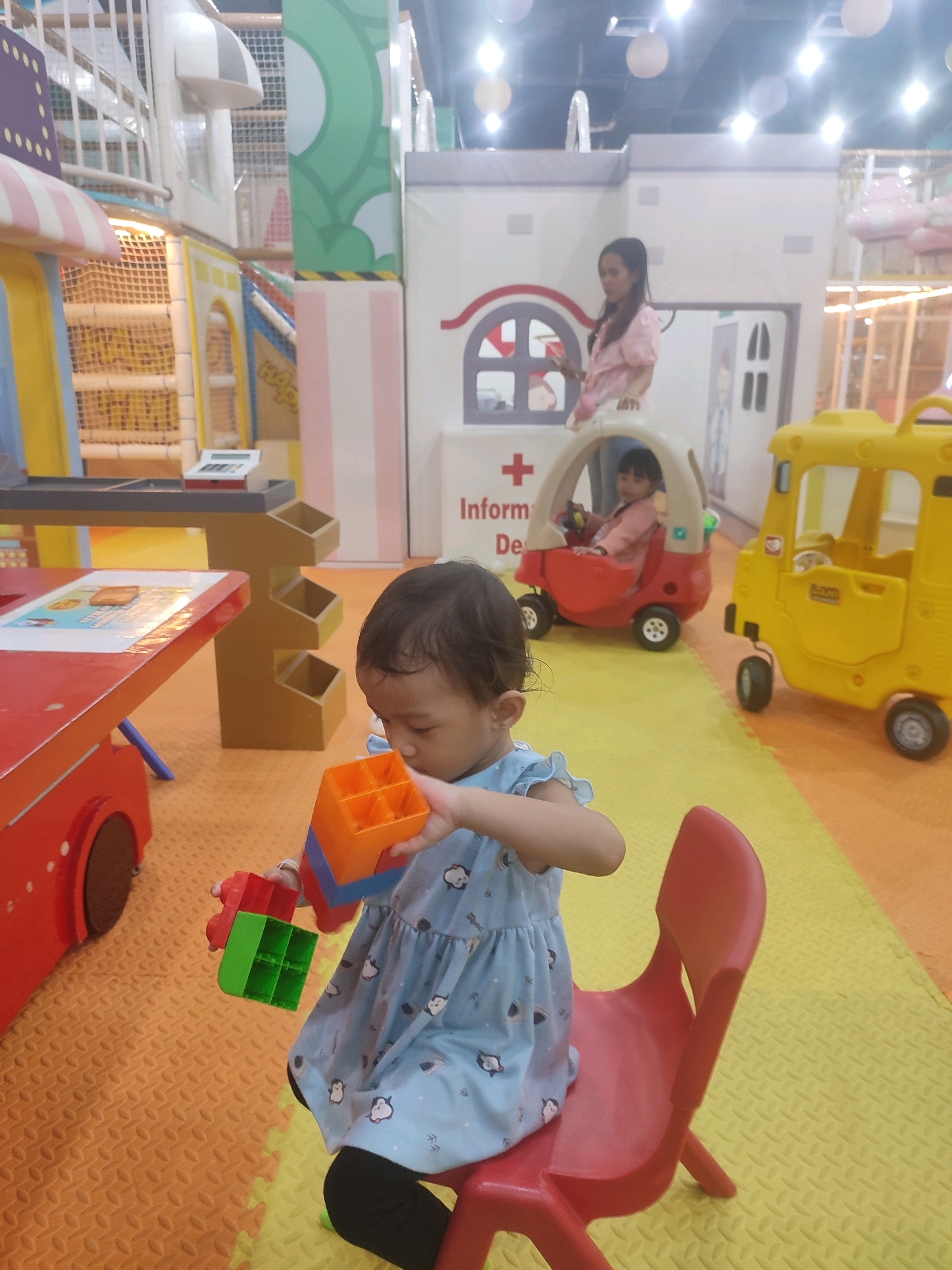 A young child wearing a light blue dress with a penguin print sits on a red plastic chair and plays with colorful toy blocks. The child is holding an orange block in their right hand and a green block in their left hand. In the background, a woman in a pink shirt stands near a toy building with a red cross symbol and text that reads "Information Desk". To the right, a yellow toy vehicle resembling a bus is parked on a yellow and orange floor mat. The scene appears to be an indoor playground or play area. The specific location in Kediri City, Indonesia cannot be determined from the image.