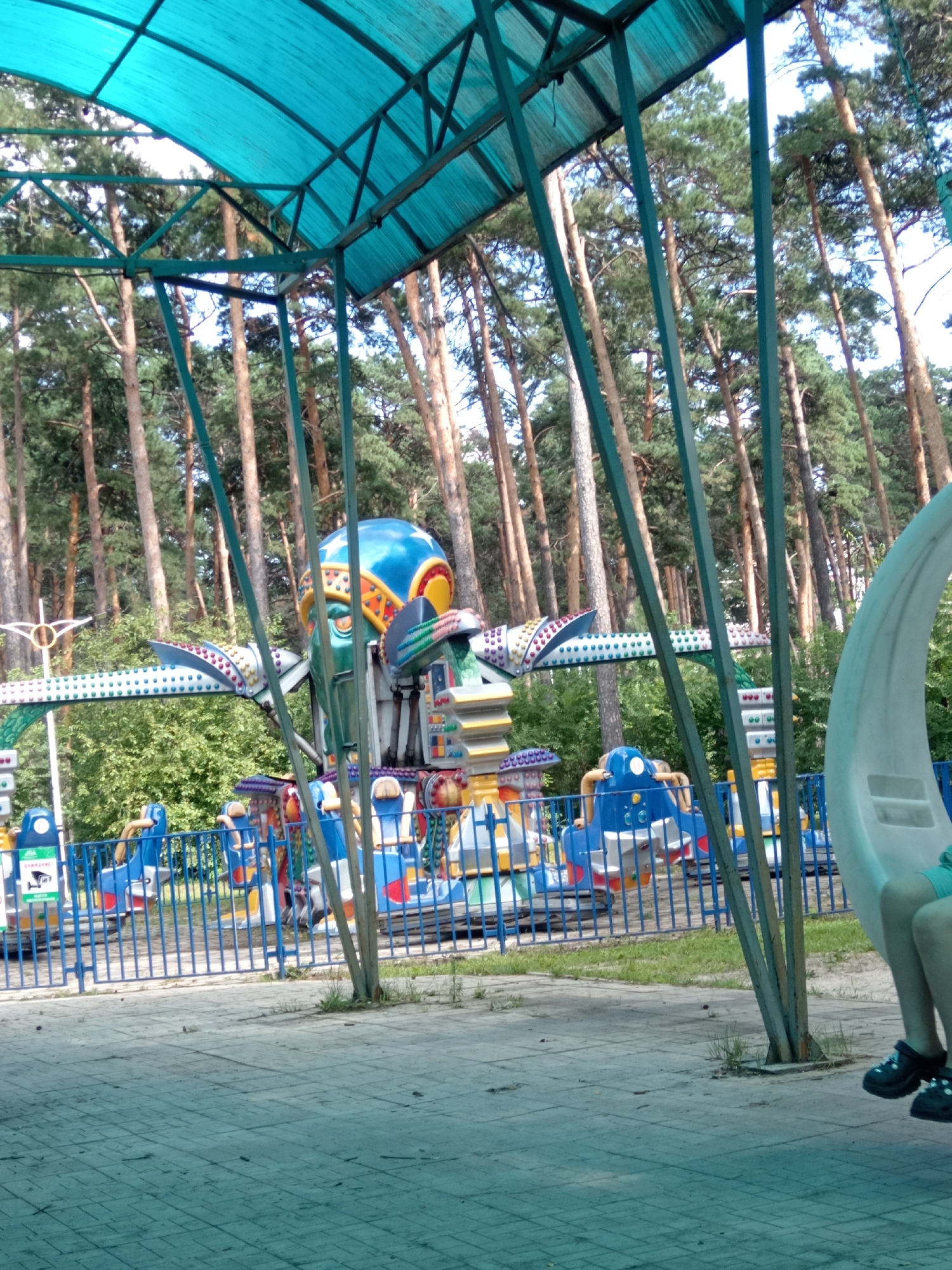 The image depicts an amusement park ride in Belogorsk, Russia. The ride features a central spinning component shaped like an alien or robot head, with multiple arms extending outwards, each holding a colorful gondola. The ride is situated amidst a forest of tall pine trees, with a bright blue canopy overhead supported by dark green metal beams. A blue metal fence encloses the ride area. In the foreground, a portion of a white crescent-shaped swing can be seen, with a person's legs and feet, wearing black shoes, resting on the paved ground below. The ground is made of gray interlocking pavers. The scene appears to be a sunny day, judging by the lighting.