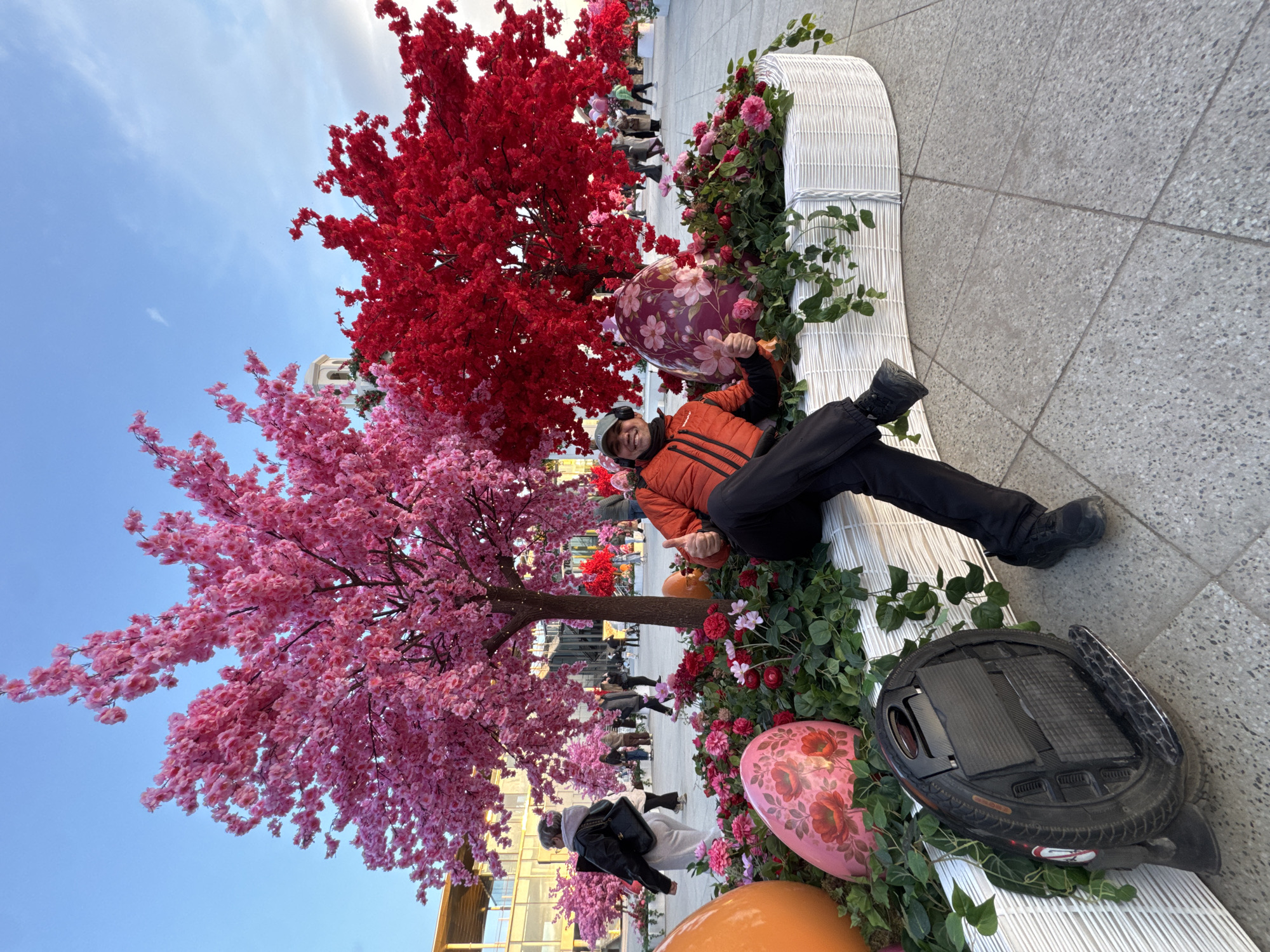 The image depicts a man lying on a white wicker structure decorated with flowers and foliage in what appears to be a public space. The man is wearing an orange jacket, black pants, and a hat. He is giving two thumbs up. Next to him is a one-wheeled electric vehicle. In the background are artificial cherry blossom trees, one with pink blossoms and the other with red. The scene includes oversized decorative eggs. Other people are visible walking in the background. Based on the architecture and overall aesthetic, the location is likely within the city of Moscow, Russia.