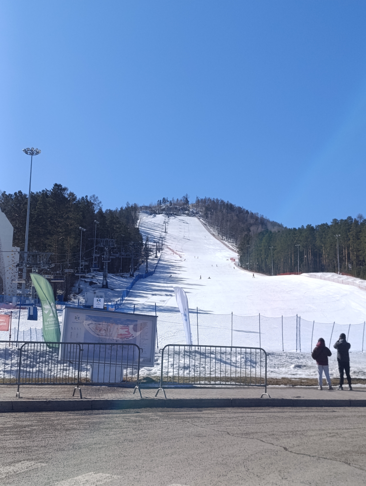 A daytime exterior shot shows a ski slope in Krasnoyarsk, Russia. Snow covers the slope, where several skiers are visible. A ski lift runs up the slope on the left. The ski area is flanked by trees on both sides. In the foreground, a metal fence runs along the edge of an asphalt road. Behind the fence are two people standing. There is a large sign depicting a map of the ski resort. Green and white flags are next to the sign. The sky is clear blue.