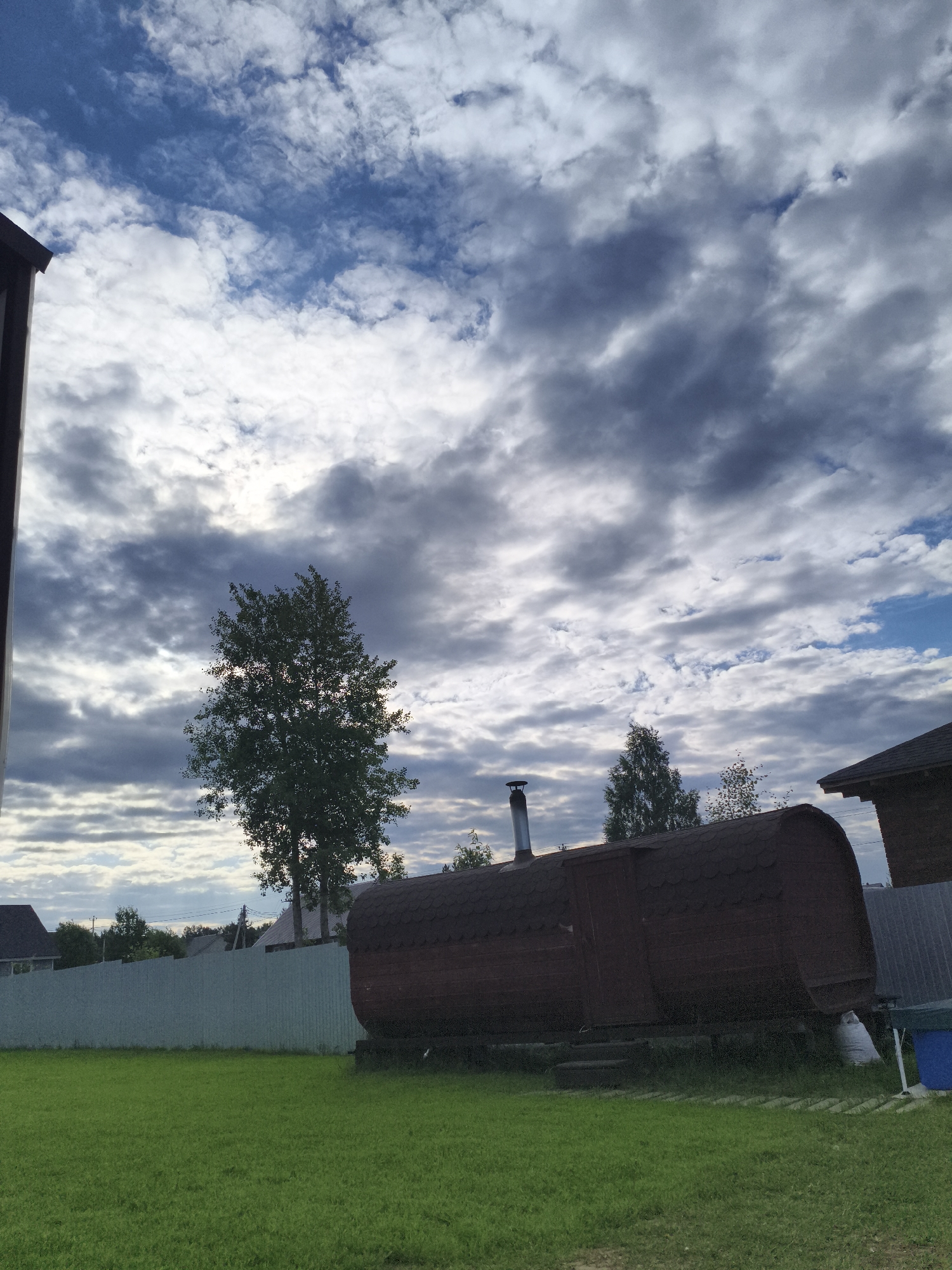 The image depicts an outdoor scene in a rural or suburban setting, likely in the evening given the dramatic cloud formations and diffused sunlight. The sky is a prominent feature, filled with a mix of dark, stormy clouds and brighter, illuminated patches suggesting either the setting or rising sun.
In the foreground, a vibrant green lawn stretches across the image. To the right, a unique, barrel-shaped structure, possibly a sauna or a small dwelling, is visible. It has a dark, shingled roof and a wooden door. A metal chimney protrudes from its top, indicating it might be functional. Next to this structure, a white bag of material is placed on the ground.
Further in the background, a pale blue corrugated fence runs horizontally, separating the lawn from more distant elements. Beyond the fence, various houses and trees are scattered, suggesting a residential area. A tall, leafy tree stands silhouetted against the sky in the middle ground, adding to the natural element of the scene. The overall impression is one of a peaceful, albeit potentially stormy, evening in a semi-rural location. No people or visible text are present in the image.