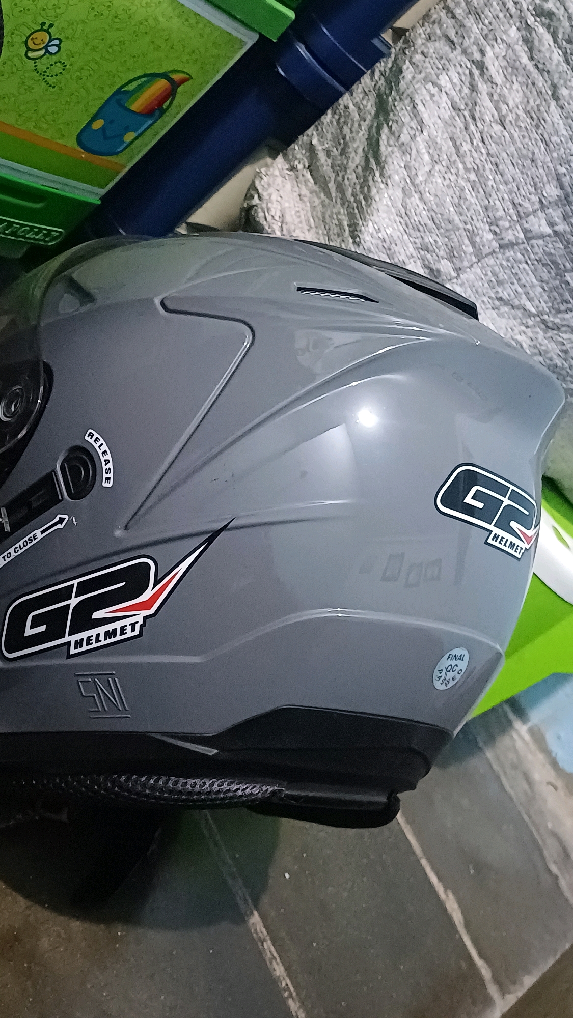 The image shows a grey helmet with "G2 HELMET" written on it in black and red letters. There is also a "SNI" sticker on the helmet. A small circular sticker with "FINAL QC PASS" is visible on the lower right side of the helmet. The helmet is lying on a tiled floor with a green object in the background. The scene is indoors, and there are no people visible. The overall lighting suggests it is daytime. There is no specific information in the image to indicate it is located in Kowangan, Indonesia.