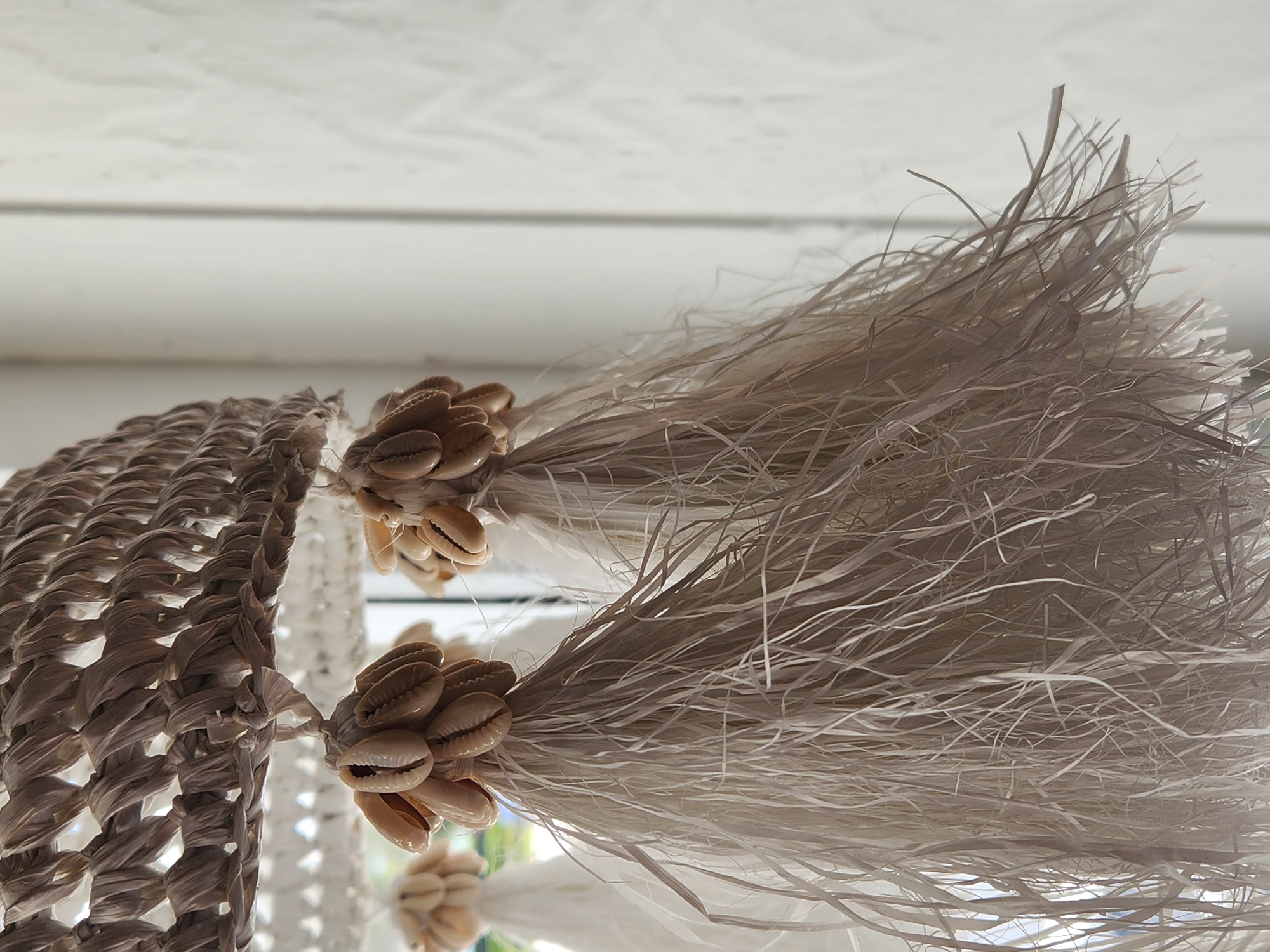 The image is a close-up of a decorative hanging light fixture with natural, textured elements. The top of the fixture appears to be woven from a light-colored, rough material. Hanging from it are three separate decorative tassels. Each tassel consists of a cluster of small, light brown seashells, likely cowrie shells, gathered at the top and bound with twine. Below the shells, the tassels are composed of long, thin strands of what looks like raffia or shredded paper, colored in shades of off-white and beige. These strands are frayed and extend downwards, creating a wispy effect. The background is softly blurred, showing a white wall with a subtle texture and the hint of natural light coming from the side. The scene evokes a coastal or bohemian aesthetic. There is no indication of a specific location within the city of Maitland, United States of America.