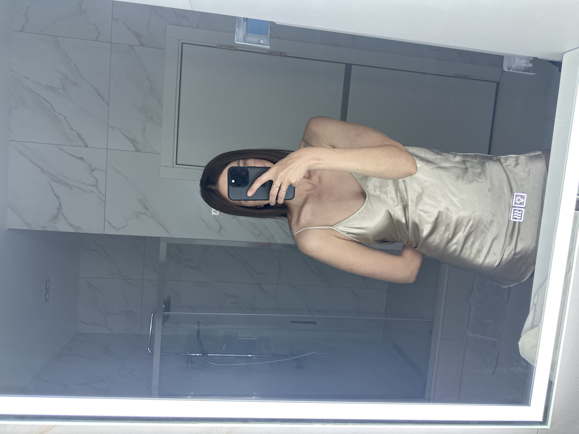 A person is reflected in a mirror, taking a selfie with a smartphone. The person, seemingly female, has shoulder-length brown hair and is wearing a light-colored satin slip dress. The setting is a bathroom with white marble-patterned tiles. The mirror reflects elements of the bathroom, including a shower with a glass door, a white cabinet above the sink, and a light fixture attached to the mirror. The location is Прикамский, Russia.