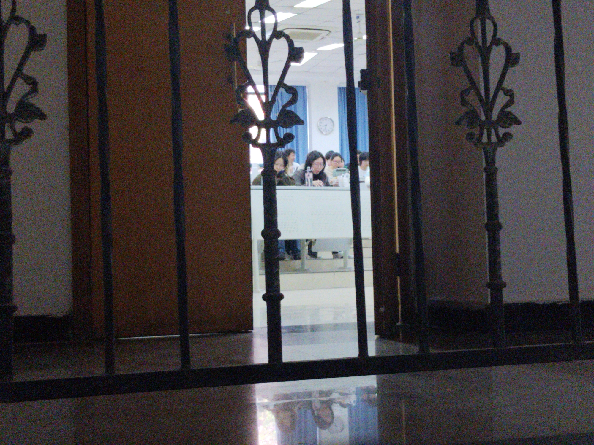 The photograph shows a group of people sitting at a table in what appears to be an office or classroom, viewed through an ornate wrought iron gate or railing. The room has blue curtains and a clock on the wall. The view is partially obstructed by a brown door. The floor in the foreground is reflective, suggesting a polished surface. The location is potentially Yuzhong District, China, based on the prompt.