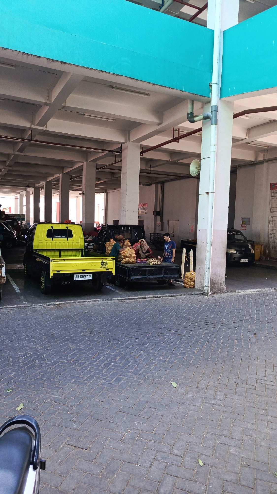 This image depicts an outdoor market scene in Ponorogo, Indonesia, likely at a traditional market or a designated area for vendors. The setting appears to be a covered market or parking area with a concrete floor and a high ceiling supported by concrete pillars. A bright teal-colored structure runs along the top of the image, possibly a roof or wall.
In the foreground, a yellow pickup truck is parked. Its bed is empty except for a yellow tarp. Next to it, a black pickup truck is parked with its bed laden with sacks of produce, likely onions or potatoes, and some root vegetables. Three individuals are interacting with the produce. One person, wearing a brown hat and green shirt, appears to be arranging the sacks. Another person, a woman in a headscarf, is seated and looking at the produce. A third person, a man in a blue shirt, stands nearby, observing.
In the background, other vehicles are visible, including a black car and what appears to be another pickup truck. The ground is paved with dark gray interlocking bricks laid in a herringbone pattern. The overall atmosphere suggests a commercial transaction or preparation for sales.