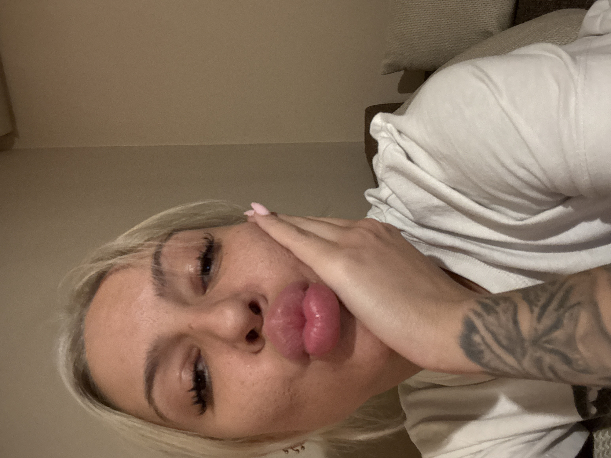 A woman with blonde hair is lying down, posing for a selfie. Her face is close to the camera and she is pursing her lips. She has long eyelashes. Her right hand is resting on her cheek. Her right arm, visible in the frame, has a tattoo. She is wearing a white shirt. The background includes a neutral-toned wall and a couch with a beige cushion. There is no evidence to place this image in Plovdiv, Bulgaria.