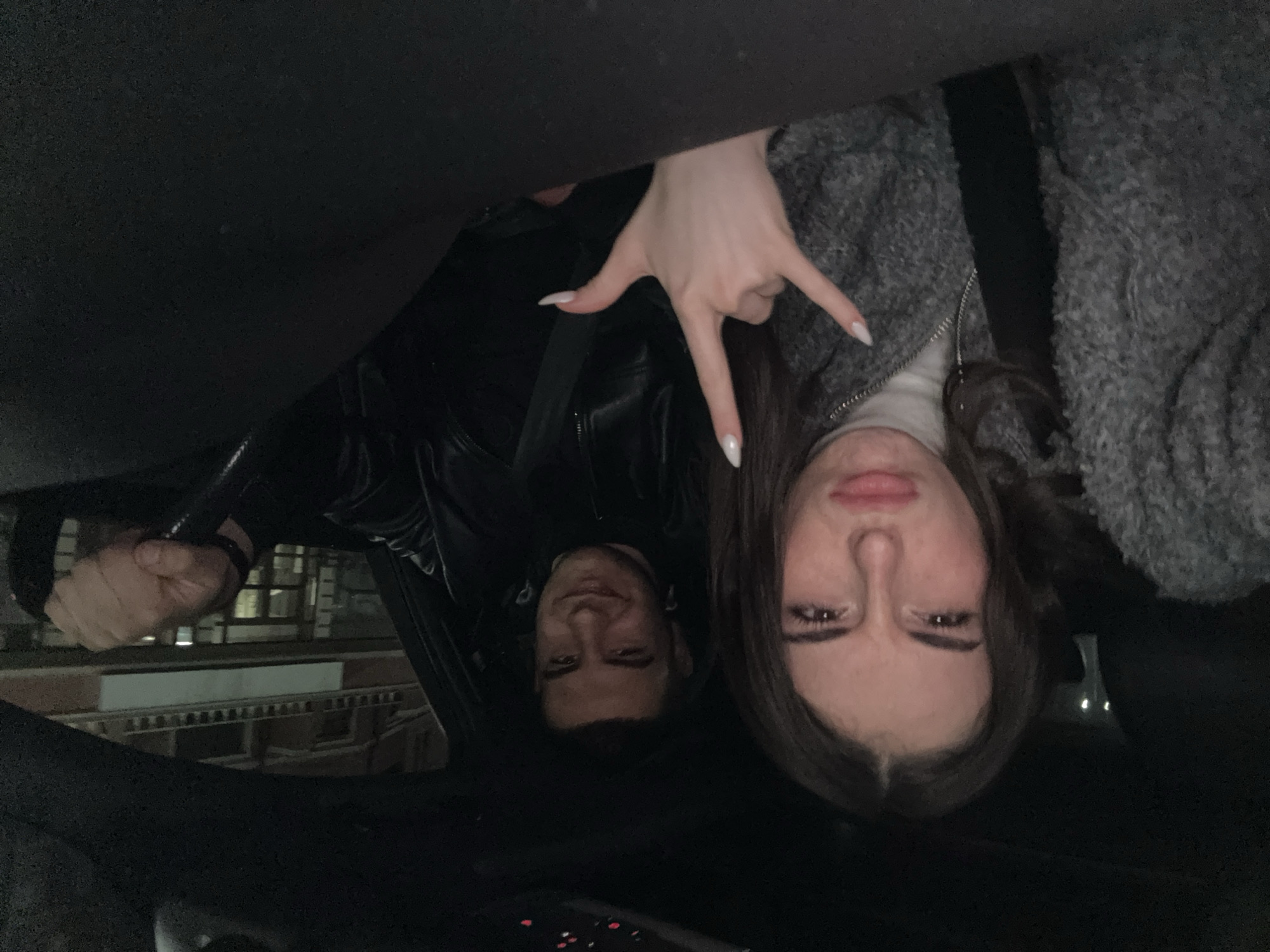 Two individuals, a male and a female, are inside a vehicle. The woman is in the foreground, with her hand raised in a "rock on" hand gesture. She wears a grey fleece jacket over a white turtleneck. Her nails are painted white. Behind her is a male, who appears to be looking toward the camera. He is wearing a black leather jacket. The scene is dimly lit, and the background shows a building exterior through the car window. Given the architecture and street layout visible through the windshield, it is potentially Plovdiv, Bulgaria. It is evening or night. The perspective indicates the photo was taken from above. The interior of the car is dark.