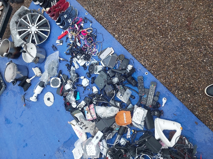This image depicts an outdoor market stall in Eckbolsheim, France. A large blue tarp is spread across a gravel or crushed stone ground surface, serving as a display for a variety of used items.
In the upper right portion of the tarp, three stainless steel pots, two of which appear to be pressure cookers, are arranged. Adjacent to these is a multi-spoke car wheel, predominantly black with silver accents. To the right of the wheel, several pairs of athletic shoes, including dark blue and red varieties, are lined up. A white cylindrical security camera and a white immersion blender are also visible in this area.
The central and left sections of the tarp are densely covered with miscellaneous electronic components and accessories. This includes extensive piles of tangled black and multicolored cables, power adapters, and numerous remote controls of various designs. Small black electronic boxes, potentially routers or media players, are scattered throughout these piles, some individually and others grouped. An orange, padded protective case is visible among the electronics. In the lower left quadrant, the hemispherical base of a white Apple iMac G4 computer is identifiable. Many smaller items, including various connectors and components, are enclosed in clear plastic bags.
At the bottom center of the frame, the toe of a human foot, wearing a dark shoe with a white sole, is partially visible, indicating a person's presence near the display. The scene is illuminated by natural daylight.