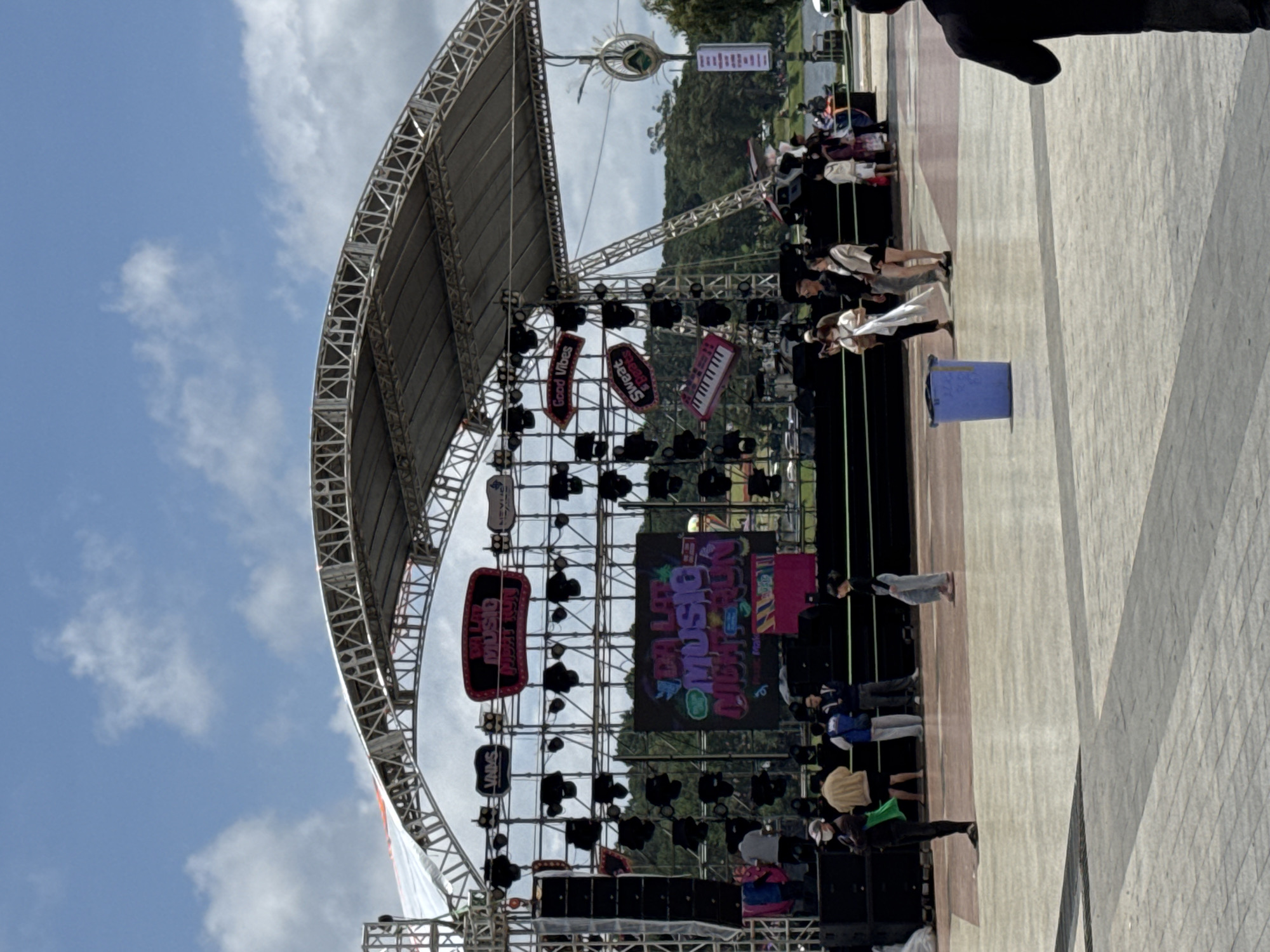 An outdoor event stage is prominently positioned under a partially cloudy blue sky in Da Lat, Vietnam. The stage structure consists of a large metal truss framework with a curved canopy of white and gray fabric. A large LED screen at the center of the stage displays "DA LAT MUSIC NIGHT RUN" in colorful, graffiti-style text. Above this screen, a red-rimmed sign reiterates "DA LAT MUSIC NIGHT RUN." Various decorative props and signs are affixed to the trussing, including "VNMS" and a stylized keyboard on the left, and "Good Vibes," "Sweat & Beats," and a large pink keyboard on the right. Multiple black stage lighting fixtures are mounted across the truss structure, and large black speaker stacks flank both sides of the stage.
In the foreground, a paved plaza area with light and dark gray tiles extends towards the stage. Approximately ten to fifteen individuals are visible in the scene. On the left side of the plaza, a person wearing a black jacket, light pants, and a white face mask stands, holding a green object. Near them, a person in a tan jacket and dark shorts is present. In the mid-ground, a blue plastic trash can sits on the paved surface. To the right of the trash can, a group of people includes a woman in a white top carrying a white bag, a man in a dark shirt, and a woman in a light jacket and dark shorts. Further right, a person in light gray pants and a black top walks away from the stage. A person in a dark suit jacket is partially visible on the far right foreground, facing away from the camera. On the stage itself, a person in a gray t-shirt and dark pants is visible on the left side, while other individuals, some wearing blue vests, are present closer to the background of the stage.
Beyond the stage, a dense treeline indicative of a forest or park is visible, and further in the background, a body of water, likely Xuan Huong Lake, can be discerned. A decorative light pole with a green sculptural element is visible on the right side of the background, adjacent to the lake. The overall lighting suggests a bright, sunny day.